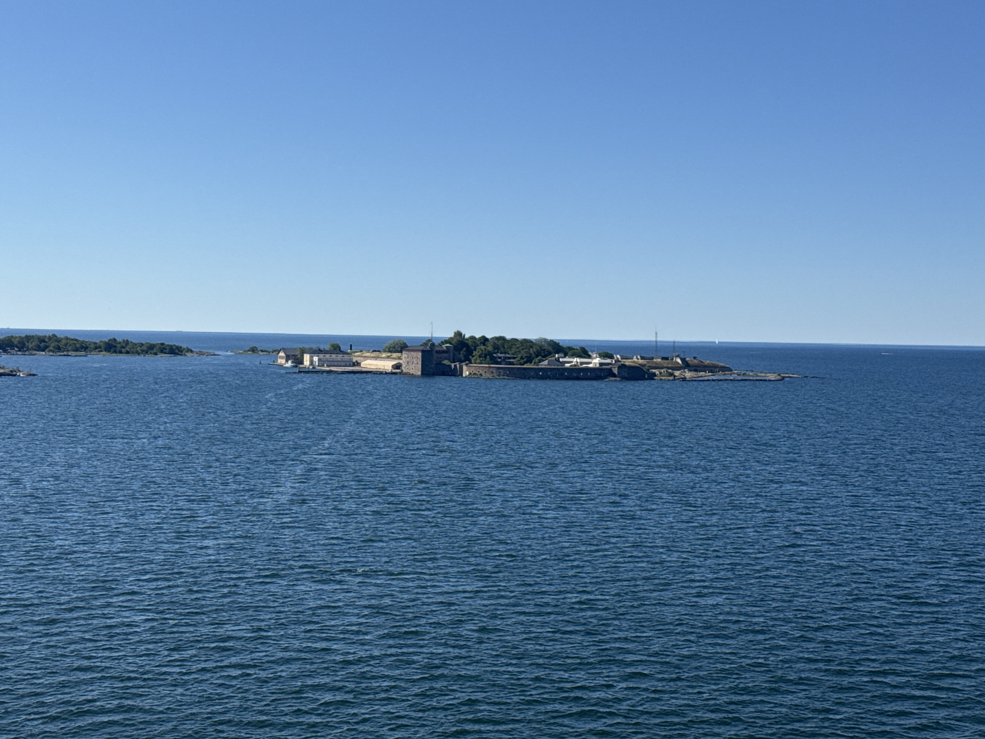 The image shows a clear blue sky and a calm blue sea. In the middle ground, there is a small island or peninsula with several buildings and fortifications. The structures appear to be made of stone and have a historical appearance, possibly part of a naval base or castle. Trees and vegetation are visible on the island. The water is rippled, suggesting a gentle breeze. The scene is devoid of people and other living beings. The location is the island of Tjärö, part of the Karlskrona archipelago in Sweden, famous for its naval heritage and fortifications.