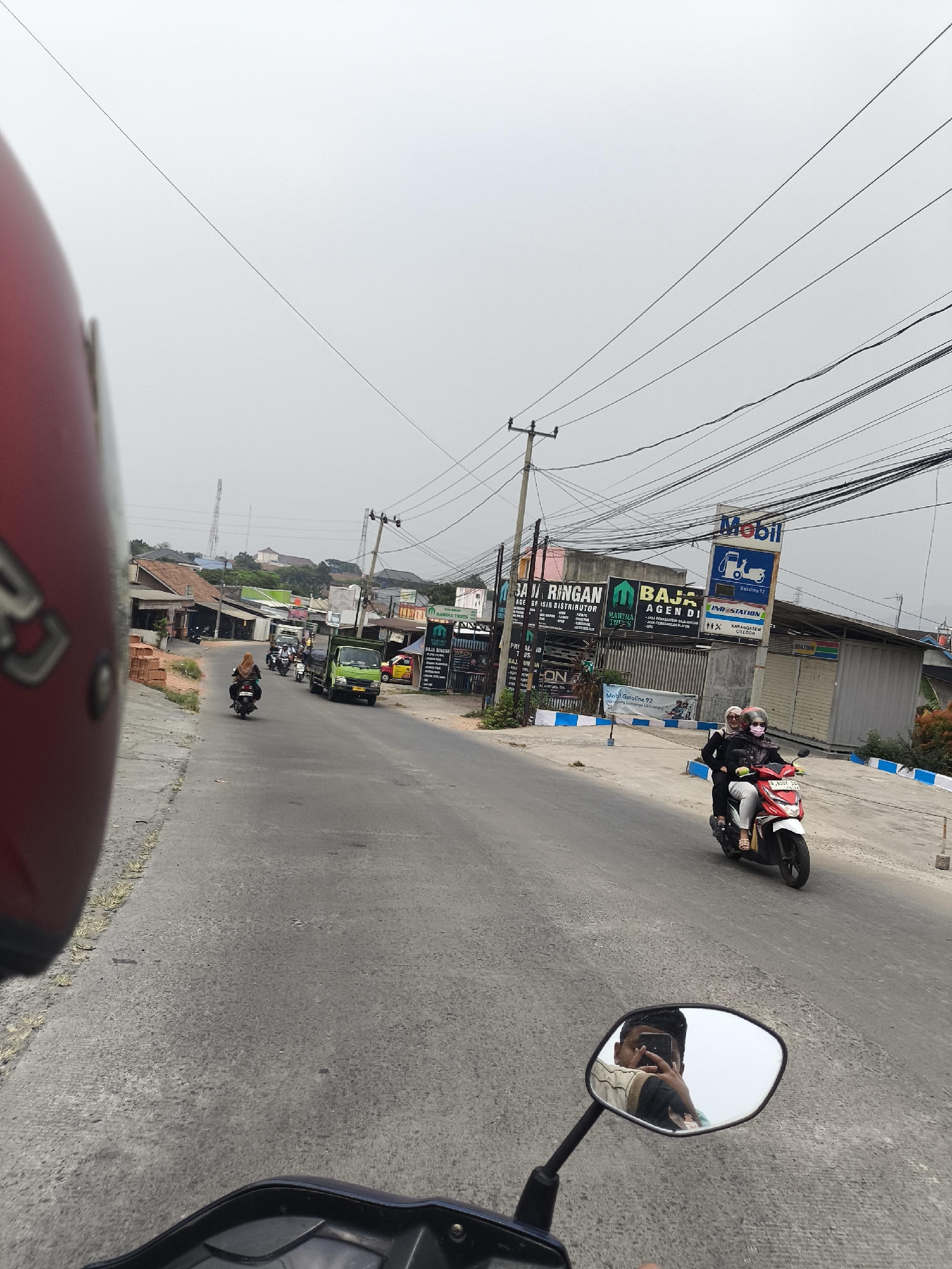 The image depicts a street scene in Cilegon, Indonesia, taken from the perspective of someone riding a motorcycle. A red helmet is visible in the foreground on the left, obscuring part of the view. The road is paved and appears to be a public thoroughfare with other vehicles present.
In the middle ground, several motorcycles are seen. One motorcycle is ridden by a person wearing a hijab and a dark top, traveling away from the camera. Further down the road, a green truck is visible, and beyond that, more motorcycles are present.
On the right side of the road, there are several buildings and signs. A prominent sign with the "Mobil" logo and "Gasoline 92" is visible, indicating a gas station. Other signs, such as "BAJA RINGAN" and "BAJA AGEN DI" are also present, suggesting commercial establishments. Two people are riding on a white and red motorcycle, traveling towards the camera. The person in front is wearing a hijab, and the person behind is wearing a pink mask.
The environment appears to be overcast, with a hazy sky. Utility poles with numerous electrical wires are strung across the scene, a common sight in urban and semi-urban areas in Indonesia. The buildings lining the street are varied in design, with some appearing to be shops or small businesses. On the far left, a pile of bricks is visible, suggesting ongoing construction or material storage. The overall impression is a busy, functional street in a moderately developed area.