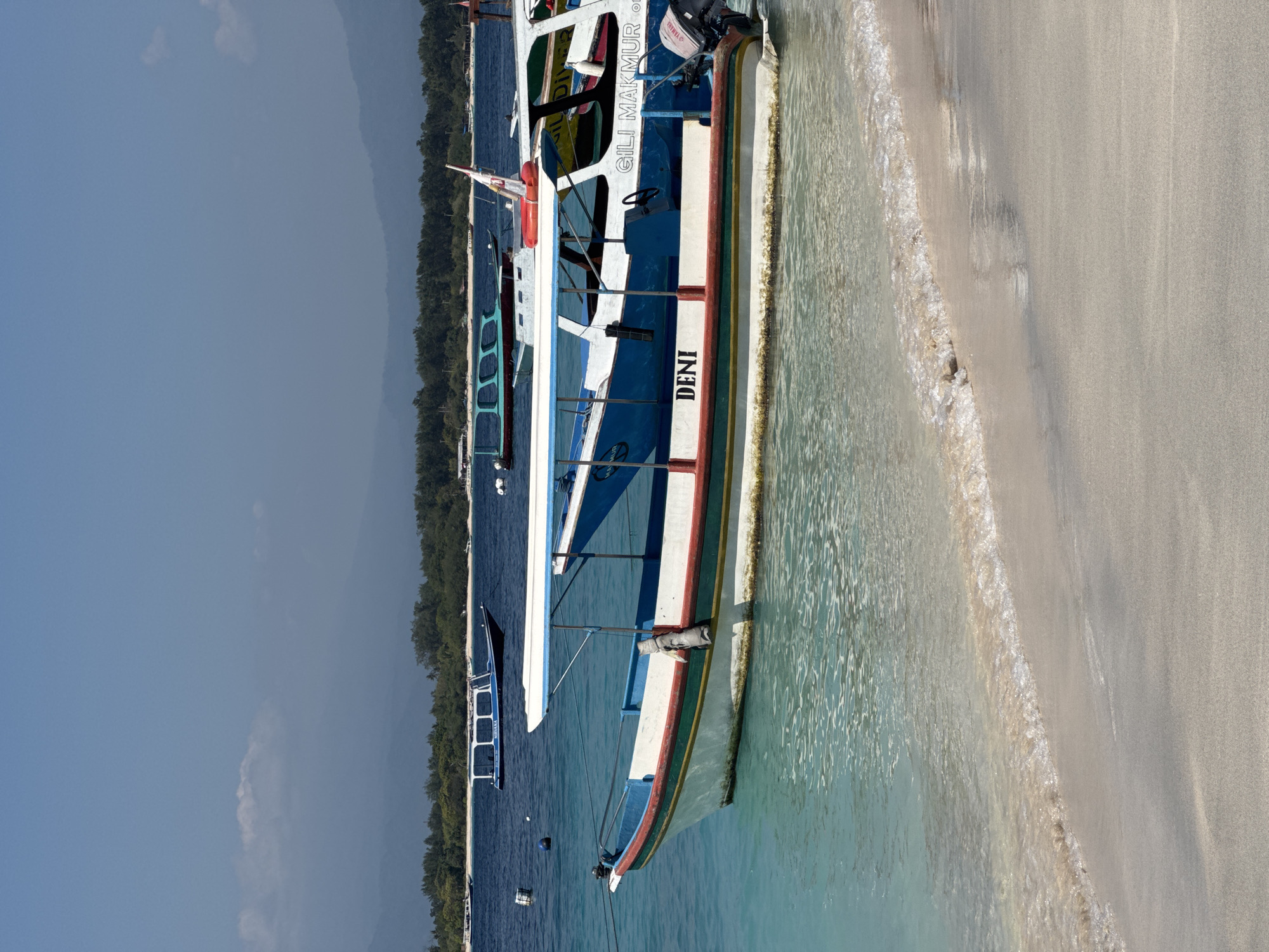 In a coastal scene, a white and blue tourist boat named "DENI" is anchored in clear, shallow turquoise water close to a sandy shore. The boat has a white canopy with blue trim and a white hull with red and green stripes. A Yamaha outboard motor is visible at the stern. In the background, a line of lush green trees and distant mountains under a hazy blue sky suggests an island or coastal landscape. Several other boats, including a blue vessel labeled "NIRVANA," are also present in the water. The tide is low, revealing the sandy bottom near the shore, with small waves gently lapping the beach. The overall impression is a serene tropical setting.