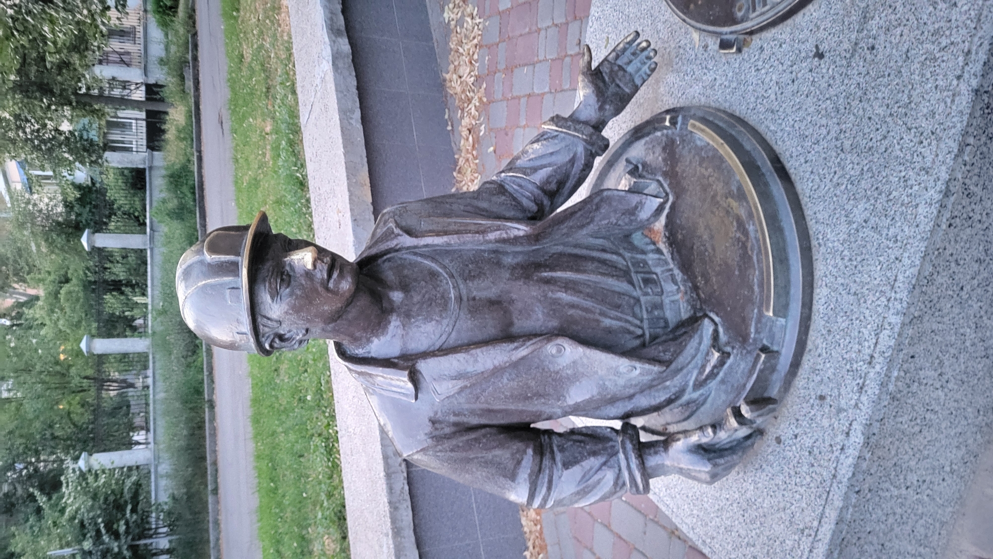 This media file shows a bronze sculpture of a man wearing a hard hat and a coat, emerging from what appears to be a manhole cover. The sculpture is positioned outdoors, with a blurred background that suggests a street or park setting with some greenery and buildings. The sculpture is made of bronze and has a weathered, metallic patina. The man's face is detailed, with his head tilted upwards and his mouth slightly open. His right arm is raised, with his hand open as if reaching out or calling for attention. His left arm is bent at the elbow, with his hand resting near his waist. The manhole cover he is emerging from is circular and appears to be made of metal, with some decorative elements. The sculpture is placed on a granite-like pedestal. The lighting suggests it is daytime, possibly overcast, as there are no strong shadows. There is no visible text in the image.
