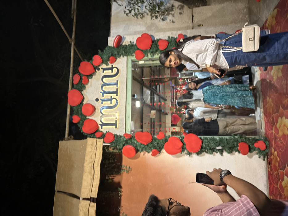 The scene depicts a nighttime outdoor view of a decorated establishment, identified by the illuminated sign "mimi" above its entrance. The entrance forms an archway heavily adorned with green foliage garlands and numerous red heart-shaped decorations of various sizes. The ground in the immediate foreground is covered by a red carpet with an orange floral pattern.
To the left, a male individual, seen from the right profile, wears glasses and a striped light-colored shirt. He holds a black smartphone horizontally, seemingly capturing a photograph or video of the decorated entrance. A wristwatch is visible on his left wrist.
To the right, a female individual stands, facing slightly towards the left but looking forward. She has dark hair and wears a light-colored, long-sleeved collared shirt and a dark denim skirt or dress. She carries a small, light-colored shoulder bag with a gold chain strap across her body and holds a smartphone in her left hand.
Inside and around the entrance, a crowd of approximately 10-15 individuals, mostly women, are visible, either entering, exiting, or congregating. One woman inside is notable for wearing a long teal-patterned skirt. The interior of the venue also features hanging red heart decorations, visible through the open doorway. To the far left, an orange-toned wall with subtle green decorative elements and an awning-like structure is visible. This scene is located in the city of Kalyan-Dombivli, India.