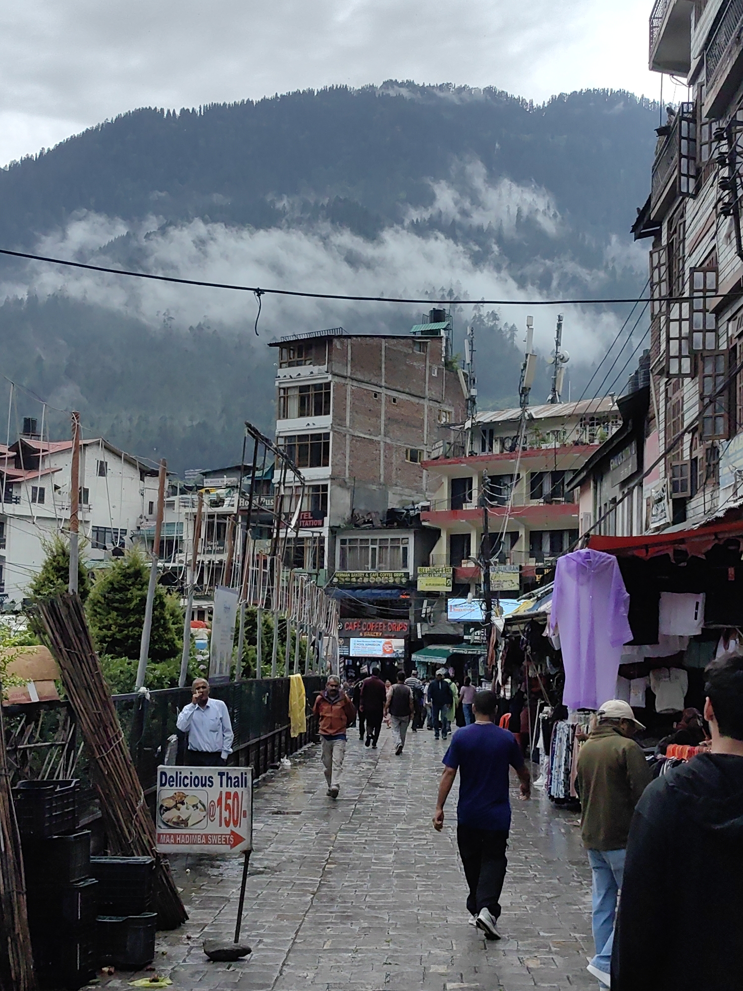 The image depicts a street scene in Manali, India, likely in the main market area, characterized by its narrow pedestrian walkway, multi-story buildings lining both sides, and a backdrop of mist-shrouded mountains. The buildings are a mix of commercial establishments and possibly guesthouses, with numerous shops displaying goods for sale, including clothing and food items. Telecommunication towers are visible on some rooftops.
The walkway is paved with stone tiles, and it appears wet, suggesting recent rain. Several people are walking along the street. In the foreground on the left, a pile of wooden poles and plastic crates are stacked. A sign advertising "Delicious Thali @ 150/-" from "Maa Hadimba Sweets" is prominently displayed.
A man in a light blue shirt and dark pants is standing near the sign, holding a phone to his ear. Further down the street, other individuals are seen walking, some browsing at shops, others seemingly heading towards or away from camera. A purple garment, possibly a raincoat or a shroud, hangs from a shop awning. The atmosphere is somewhat overcast, contributing to the misty appearance of the distant mountains covered in dense pine trees. The overall impression is that of a busy street in a hilly tourist town.