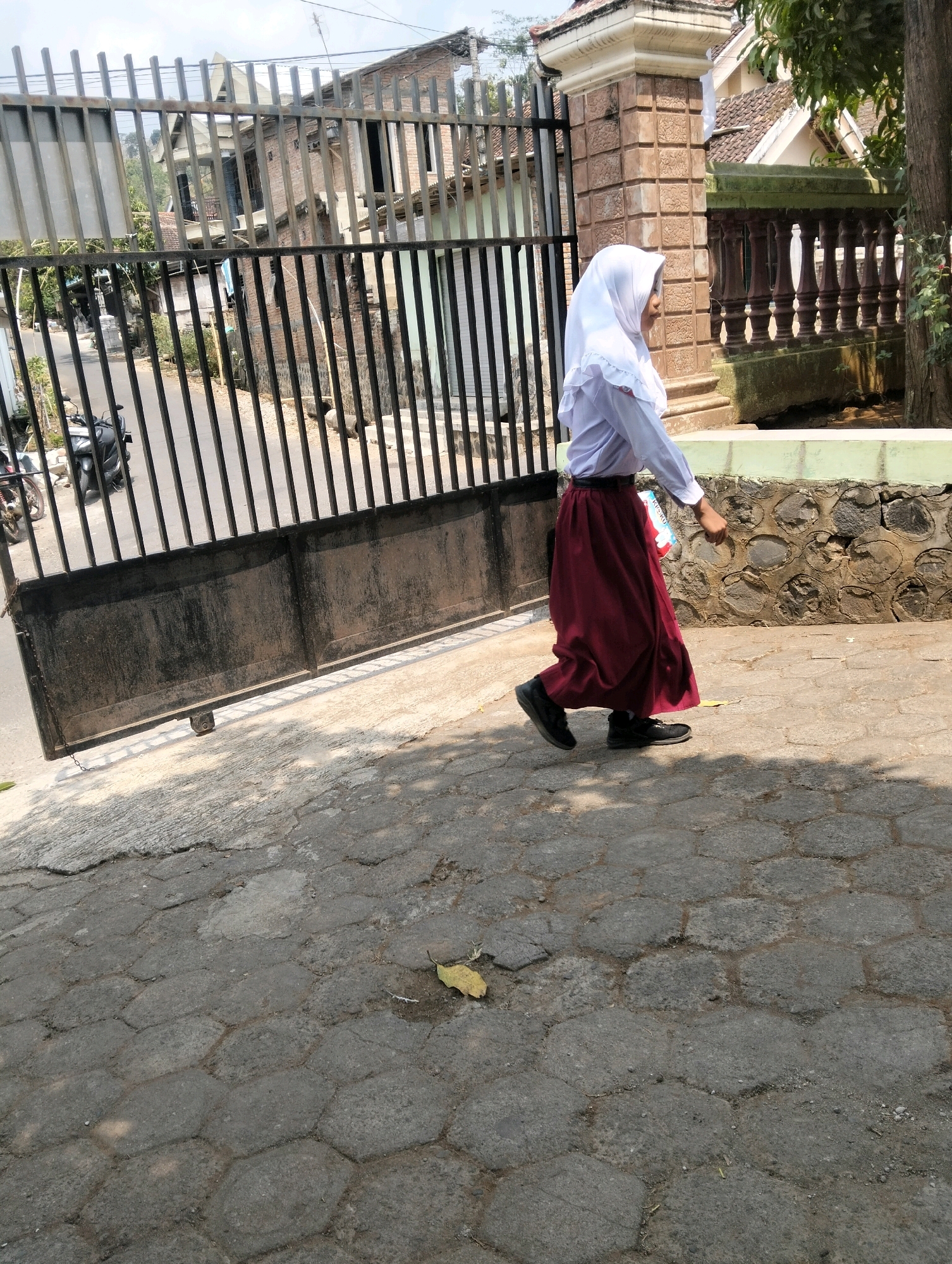 A young girl wearing a white hijab, a light blue long-sleeved shirt, and a maroon skirt walks on a hexagonal-patterned paved sidewalk in front of a black metal gate. She is walking from left to right and holding a red and white book. The gate is slightly ajar, revealing a street with a motorcycle parked on the left. Behind the gate, buildings with brick and light-colored walls and tiled roofs are visible. To the right of the gate, there is a decorative stone pillar and a stone wall. Trees and foliage are present in the background. The scene appears to be a residential area in Kanyoran, Indonesia, on a sunny day, casting shadows on the ground.