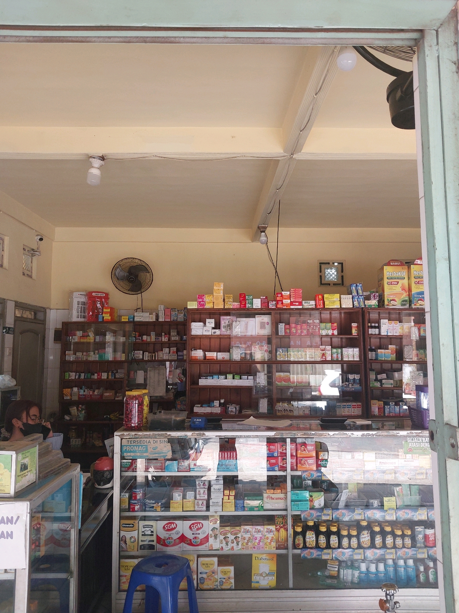 A pharmacy interior is depicted, likely in Malang, Indonesia, due to the presence of Indonesian text on signage and product packaging. The scene is characterized by wooden shelving units filled with various medicines, health products, and over-the-counter remedies. The lighting is fluorescent and the overall impression is one of a functional, somewhat dated, retail space.
In the foreground, a blue plastic stool is placed in front of a glass display cabinet. This cabinet showcases rows of boxed medications, bottles, and other health-related items. Some prominent brands and product types visible include "SGM" (a milk formula), "Tropicana Slim" (a diet product), "Listerine" (mouthwash), and various pharmaceutical boxes with Indonesian labels. A woman wearing glasses and a black face mask is partially visible in the lower left corner, appearing to be interacting with a counter or shelf. She is looking towards the left.
Behind the display cabinet, more shelves are visible, stocked with a diverse range of products. These include jars of colorful candies or supplements, boxes of medicine, and other packaged goods. A black oscillating fan is mounted on the wall above the shelves.
The walls are painted a pale yellow, and the ceiling is a lighter, creamy color. Two light fixtures are visible on the ceiling, one a simple bulb holder and another a more enclosed fixture. A small security camera is mounted on the upper left wall. The entrance to the pharmacy is partially open, revealing a glimpse of the outdoors or an adjacent area. The overall environment suggests a typical neighborhood pharmacy.
