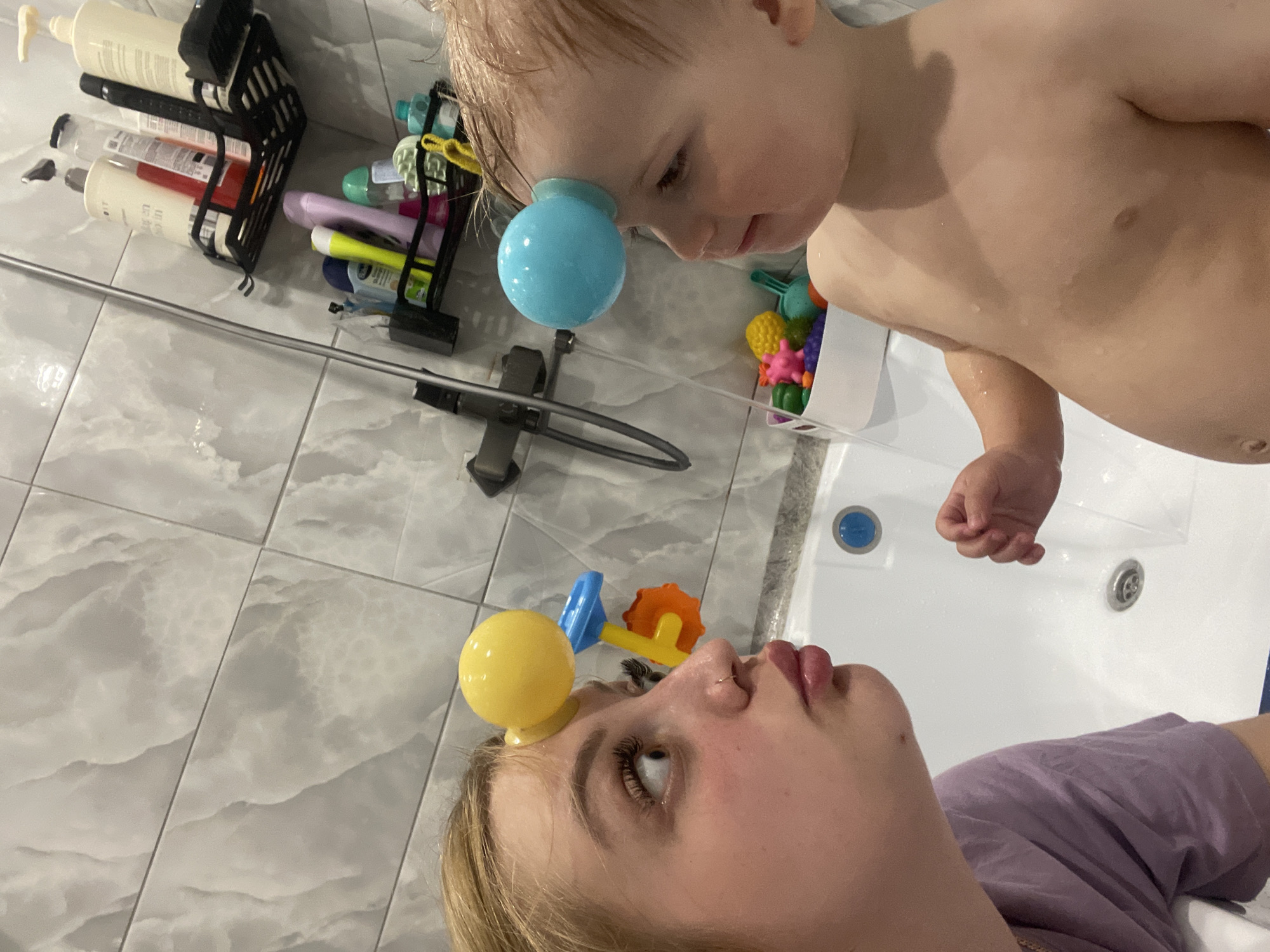 A selfie shows a woman and a young child in a bathroom. The woman, wearing a purple shirt, looks up at the child. The child, shirtless and wet, looks down at the woman. A blue suction cup toy is stuck to the child's forehead, and a yellow suction cup toy is stuck to the woman's forehead. The background includes gray tiled walls, a shower caddy with bottles, and a white bathtub. The location is not specified.