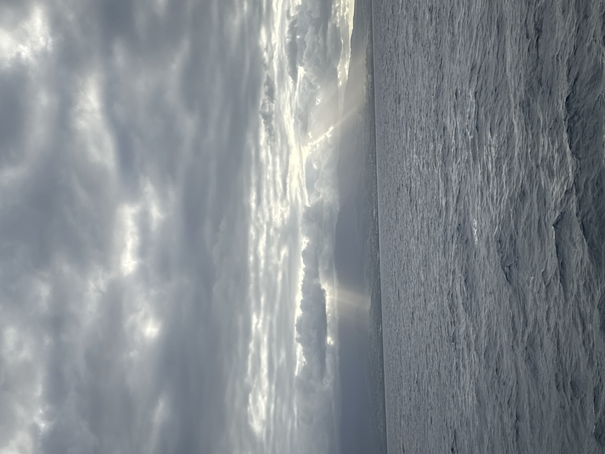 A vertical, medium shot captures a wide expanse of the ocean meeting a distant coastline under a dramatic, cloudy sky. The sea, in the foreground and taking up the lower half of the frame, is a choppy, dark blue-gray, with visible waves and ripples. Beyond the sea, a long, low-lying coastline stretches across the horizon, partially obscured by atmospheric haze and fog. The landmass appears to be mountainous or hilly, with faint outlines of structures visible along the shore.
The sky above is filled with heavy, dark gray clouds, but shafts of light are breaking through in several places, creating a dramatic effect. Most prominently, a set of sunbeams pierces through a gap in the clouds over the coastline, illuminating a portion of the land with a bright, almost ethereal glow. The overall mood is somber and majestic, evoking a sense of natural power and the vastness of the ocean. The location is identified as Capesterre-Belle-Eau, Guadeloupe. There are no people, animals, or specific objects in the image, only the elements of nature: water, land, and sky.