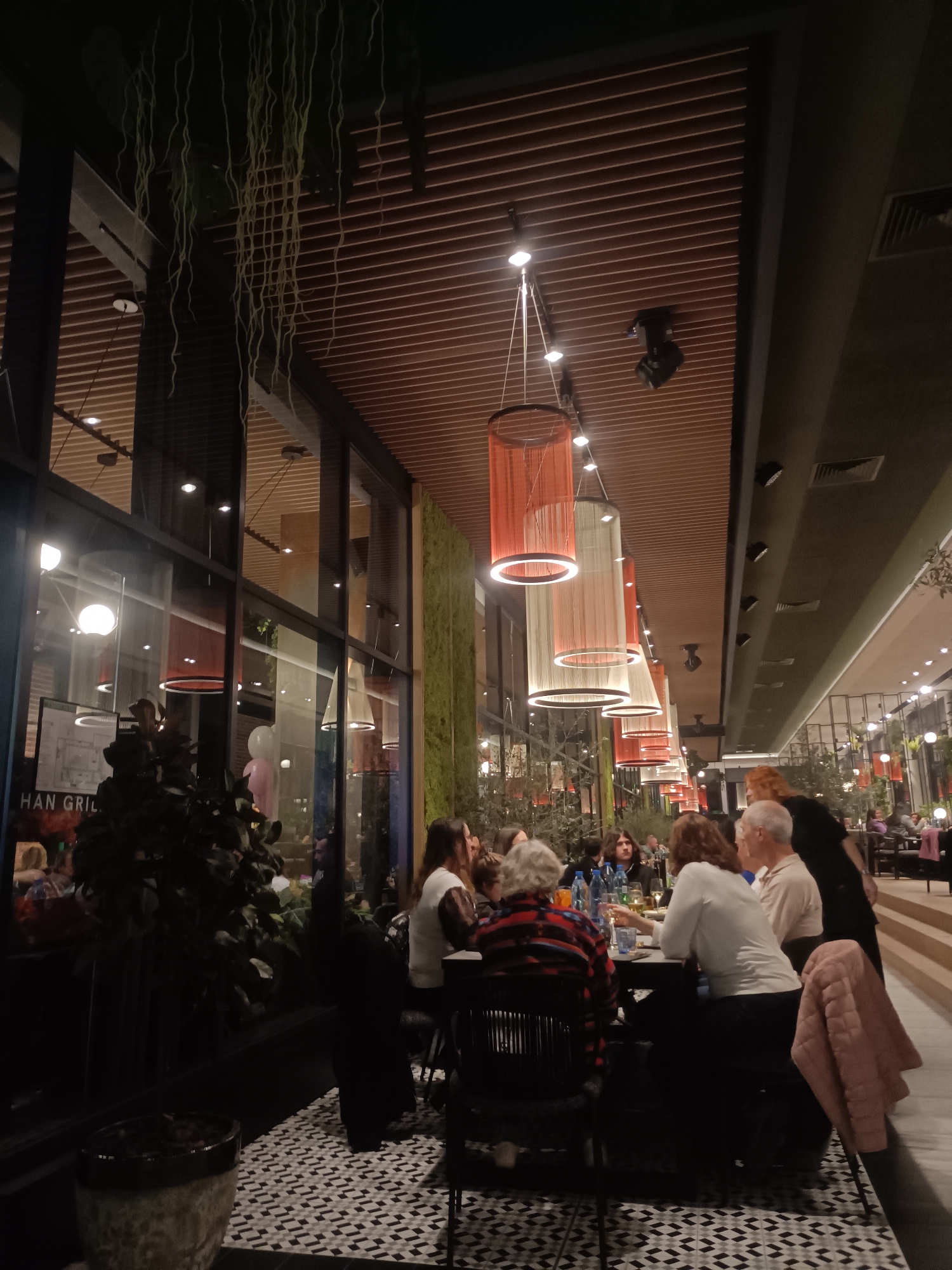 An indoor restaurant scene is depicted, likely in the evening, located in Plovdiv, Bulgaria. The image focuses on a dining area with a high ceiling featuring parallel wooden slats and a row of cylindrical pendant lights, alternately orange and white, providing primary illumination. Recessed spotlights are also visible.
In the midground, a group of approximately eight adults is seated around a dark rectangular table. One individual, wearing a red and black patterned shirt, faces away from the camera. Another, in a white long-sleeved shirt, faces the table, holding a glass. A bald man in a beige shirt sits to their left. A woman with reddish-brown hair, dressed in a dark top, stands near the table's edge on the right. A light pink quilted jacket is draped over a chair on the far right. Multiple blue water bottles and glasses are visible on the table.
To the left, large floor-to-ceiling windows reflect interior lighting and display partial text "HAN GRIL." A vertical panel covered in green moss-like material is situated between the window section and the dining tables. Dark potted plants are positioned in the foreground and near the windows. From the ceiling on the left, numerous long, thin, light-colored hanging decorations or artificial vines descend. The floor beneath the tables is covered in a geometric pattern of black, white, and gray tiles. In the background, additional patrons are visible at other tables, some situated on a slightly raised platform. The overall environment suggests a modern, spacious dining establishment.