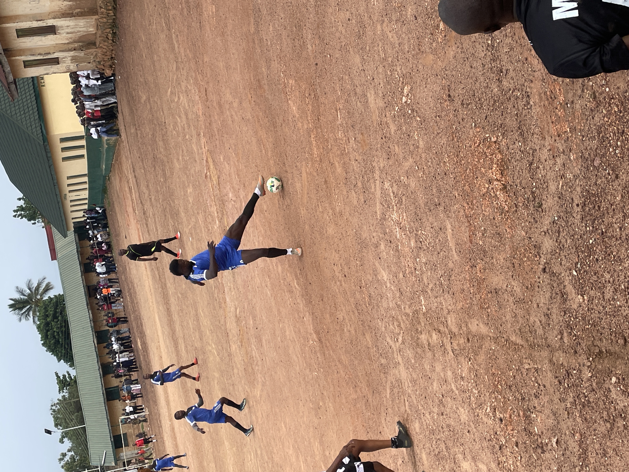 An outdoor, eye-level, medium shot depicts a football match on a dirt field in Jos, Nigeria. The field is reddish-brown and unevenly packed, with scattered pebbles and dust. In the foreground, a person in black shorts and black socks is partially visible, facing away from the camera. To the left, a person in blue shorts and a blue jersey is running, with their left leg extended as if kicking. Another person in blue shorts and a blue jersey is in the mid-ground, facing right and appearing to have just kicked a football. The football itself is visible near the center of the frame. In the background, a long, single-story building with a green corrugated metal roof stands parallel to the field. The building has a row of windows and what appears to be a sign reading "R.C.M." above some of the windows. Spectators, dressed in casual attire, are gathered in clusters along the building's edge and further back. Several individuals are standing or sitting near the building. A tall palm tree is visible behind the building, and other trees and vegetation are present in the distance. The sky is overcast with a pale, hazy appearance. The overall scene suggests a local sporting event taking place in an urban or semi-urban setting.