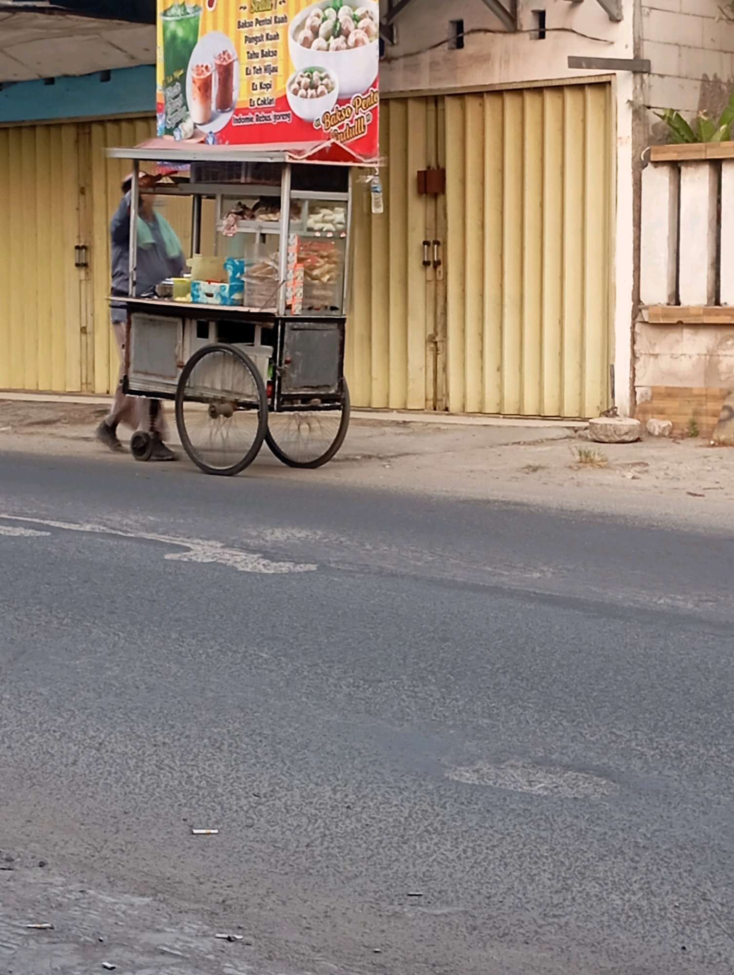 A street food cart, displaying various drinks and bowls of food, is parked on the side of a paved road. A person, wearing a dark shirt and light-colored pants, is standing next to the cart, appearing to be attending to it. The cart has large wheels and is laden with items, some of which are visible in clear containers. Behind the cart is a building with a large, yellow, corrugated metal roll-up door. To the right of the door, there is a section of brick wall. The scene is consistent with a street vendor operating in an urban or semi-urban environment in Babelan, Indonesia.
