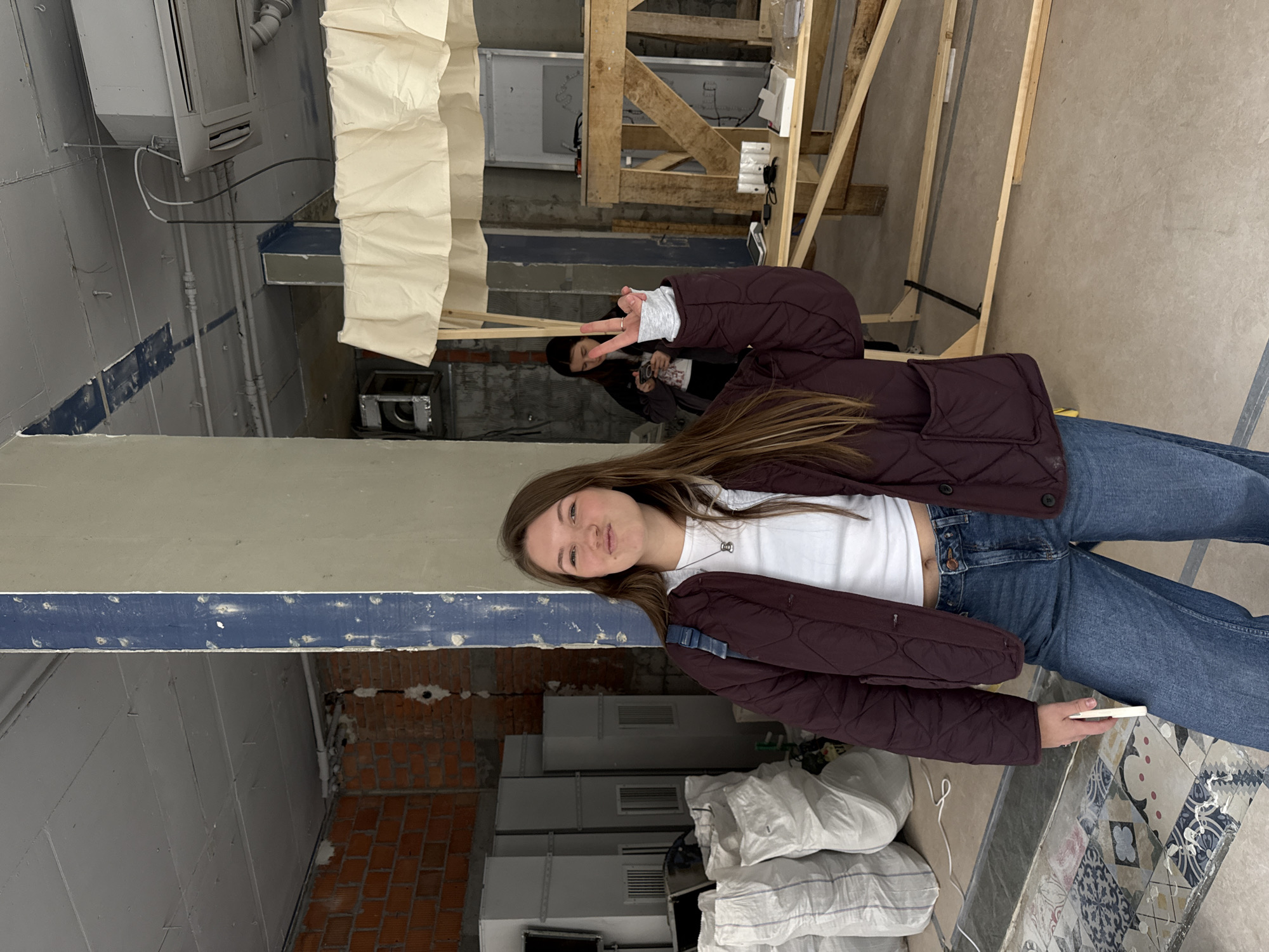 A young woman with long brown hair stands in a room under construction. She wears a white t-shirt, blue jeans, and a burgundy quilted jacket. She is facing the camera and slightly smiling. In her right hand, she holds a white object. Behind her is a person partially visible making a peace sign. The background includes exposed brick walls, construction materials (lumber, white bags filled with an unknown substance, air conditioning units), exposed wiring, and partially finished walls with blue trim. The location is possibly in Belgrade, Serbia.