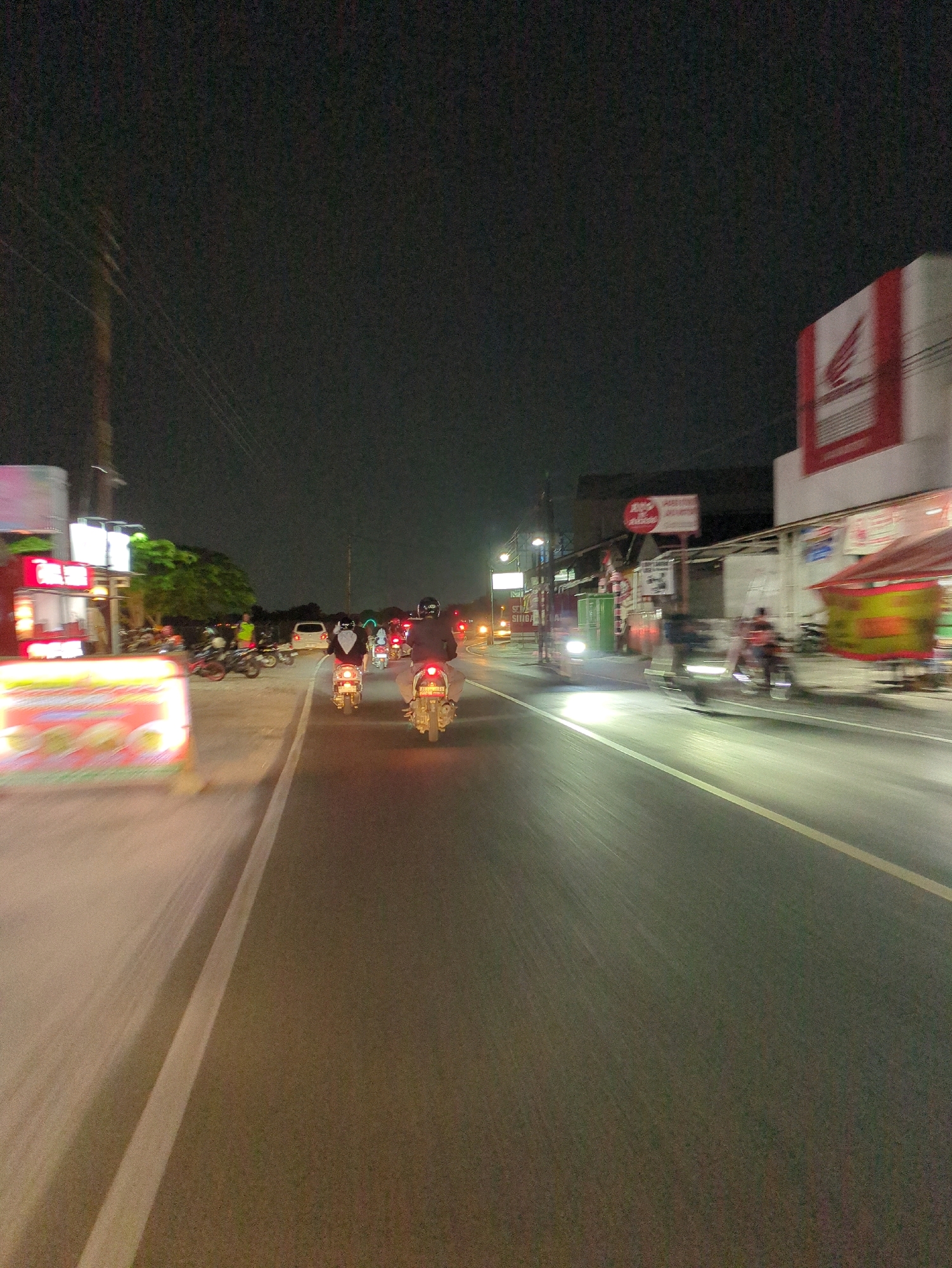 The image depicts a night scene on a road in Desa Ngesrep, Indonesia. Several motorcycles are visible, some moving forward, indicating traffic. A white car is also present. On the right side of the road, a building with a red Honda logo is prominent. To the left, there are various stalls or shops, one of which has a brightly lit sign displaying colorful images, possibly advertisements for food. Streetlights and the headlights of vehicles illuminate the scene. The overall impression is one of a busy street at night, likely in a commercial or residential area of the village.