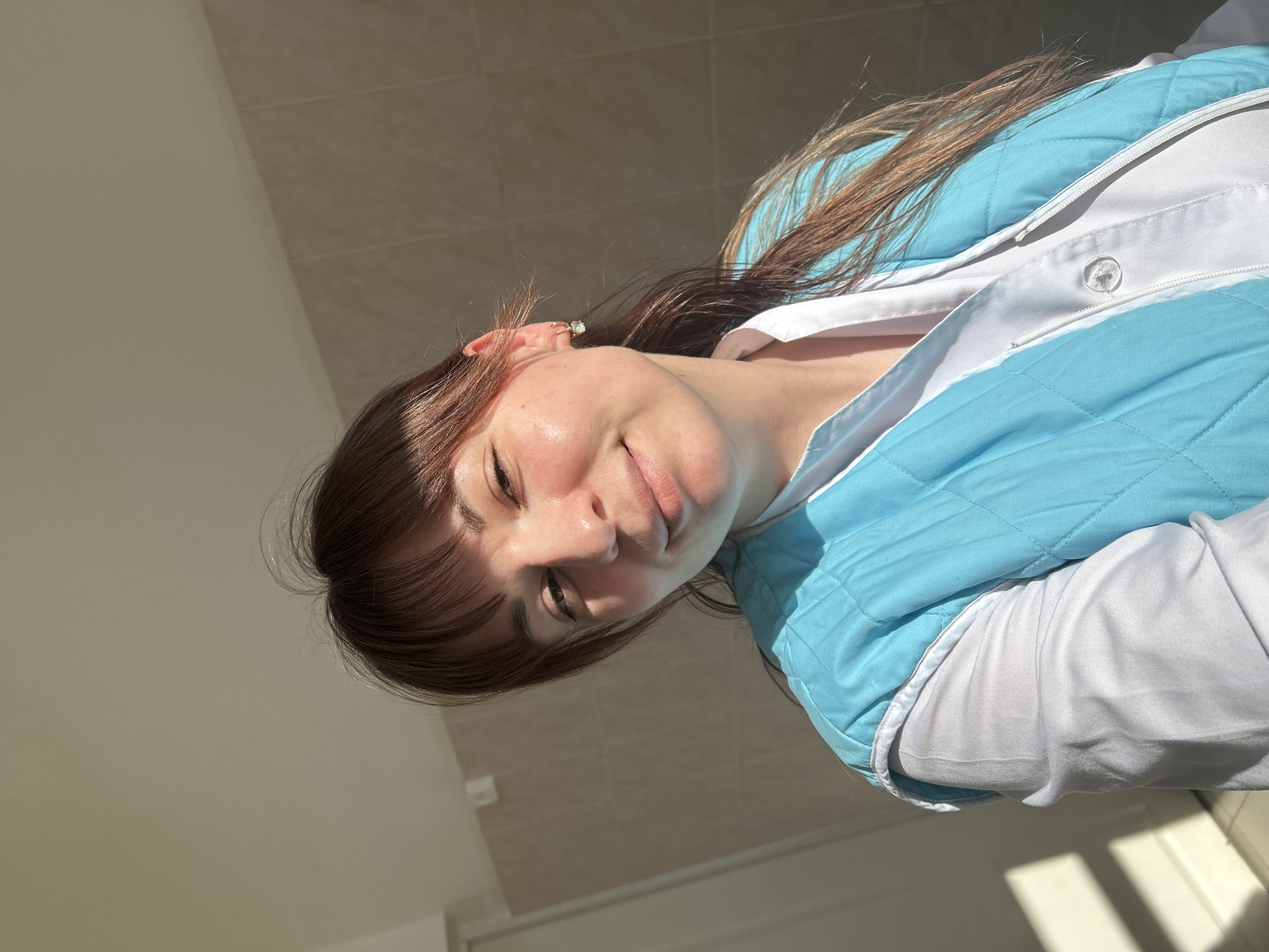 An adult female is depicted in a close-up, chest-up portrait, facing forward with a faint smile. She has light brown hair with bangs across her forehead and longer strands flowing over her shoulders. Her visible right ear features a small, gold-colored hoop earring with a clear or light-colored gem. She is dressed in a white long-sleeved shirt or uniform top, over which she wears a light blue, quilted sleeveless vest with white trim. The vest exhibits a diamond stitching pattern. The setting appears to be indoors, against a background of light-colored, possibly beige, square or rectangular tiled walls with visible grout lines, topped by a plain lighter wall or ceiling. Strong, direct sunlight illuminates the scene from the right, casting highlights on the subject and shadows on the background. The specific location in the city of Фоки, Russia, cannot be confirmed from the visual evidence provided in the image.
