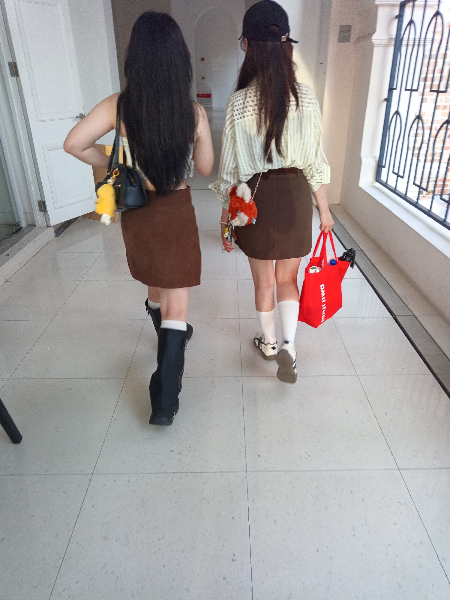 Two individuals, viewed from behind, walk down a tiled hallway. The individual on the left has long black hair, wears a brown skirt, black boots, and carries a black handbag with a yellow charm. The individual on the right wears a striped shirt, a brown skirt, white socks, sneakers, and carries a red tote bag. The hallway features white doors and a black metal grate on the right. This scene is located in Kuala Lumpur, Malaysia, though the specific building or area is not identifiable from the provided image.