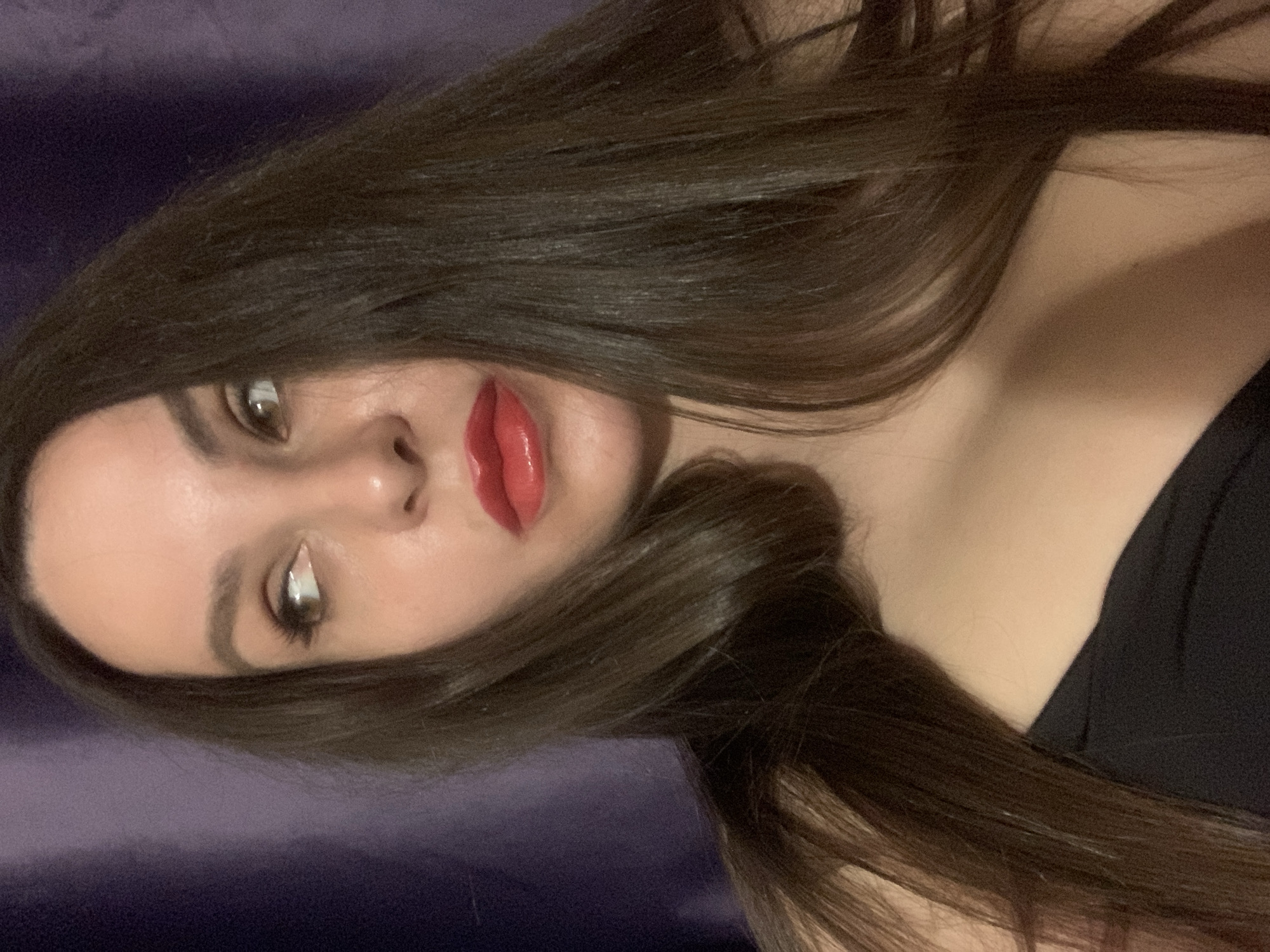 An adult female is captured in a close-up portrait, set in the city of Unknown, Unknown. She possesses long, straight, dark brown hair that cascades over her shoulders. Her full lips are adorned with bright, glossy red lipstick. Her brown eyes are directed sharply to the right side of the frame, with her head subtly tilted towards her left. Her right eye is fully visible, while her left eye is partially obscured by her hair and angle. She exhibits well-defined eyeliner and mascara, complementing her dark, arched eyebrows. A black garment, likely a top, is visible on her upper torso. The background consists of a solid, dark purple or indigo textile, displaying subtle vertical folds consistent with a curtain or draped fabric. Soft, directional lighting illuminates her face from the front, casting gentle highlights on her skin and the reflective surface of her lips. The overall lighting suggests an indoor, dimly lit environment.