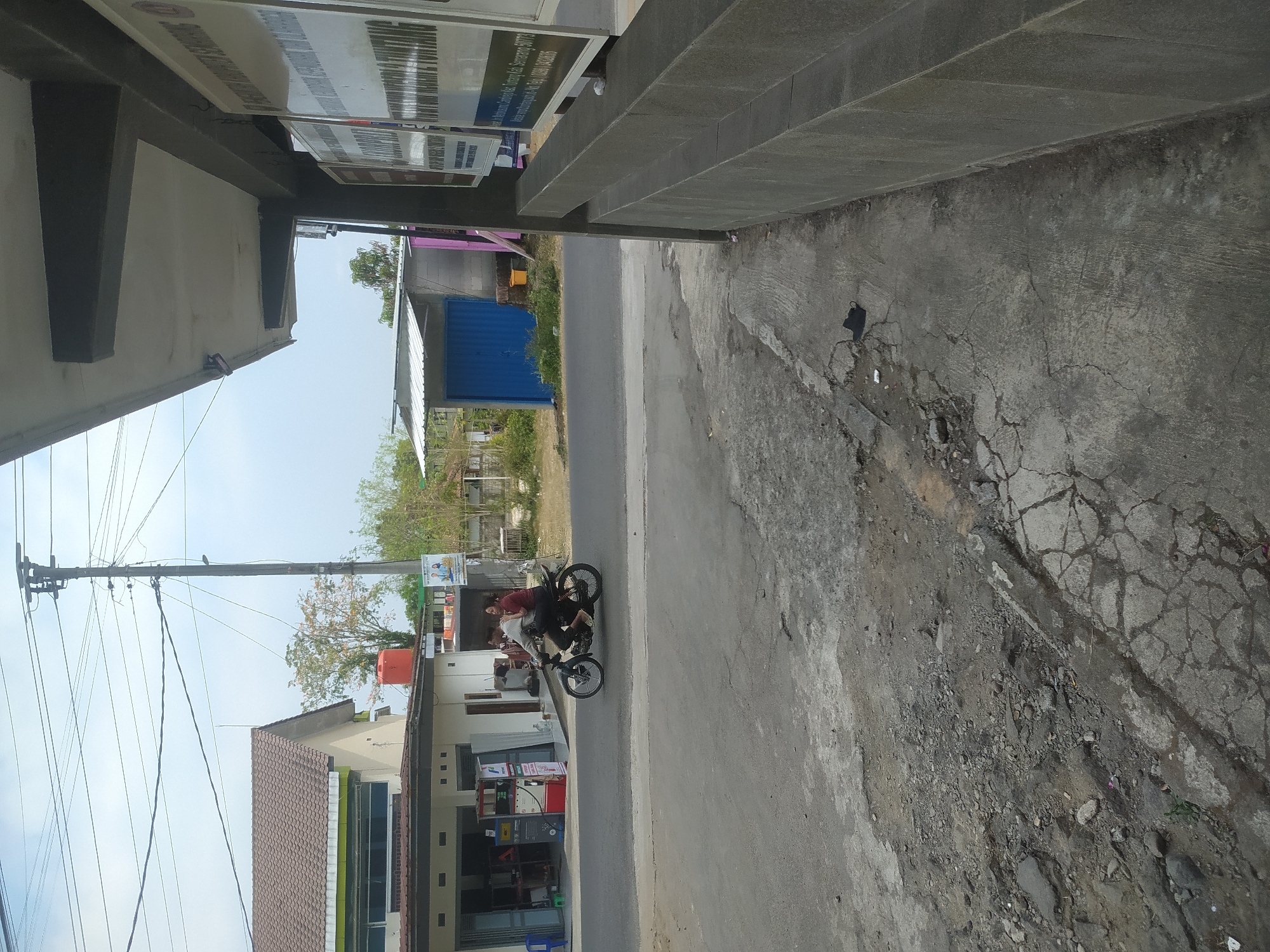 The image depicts a street scene in Candirejo, Indonesia, on a bright, overcast day. In the foreground, a cracked asphalt road with patches of loose gravel and debris leads into the middle ground. To the left, a building with a red-tiled roof and white walls has a row of storefronts, including a gas station with visible pumps. Two individuals are riding a motorcycle on the road, with the driver in a light-colored shirt and the passenger in a darker top. Beyond the building, a blue corrugated metal structure stands next to other low-rise buildings. In the background, trees are visible, and utility poles with numerous wires stretch across the sky. To the right of the scene, the corner of a modern building with a gray, textured facade and overhang is present, displaying several framed signs with text. The overall impression is one of a functional, semi-urban environment with elements of commercial activity and infrastructure.