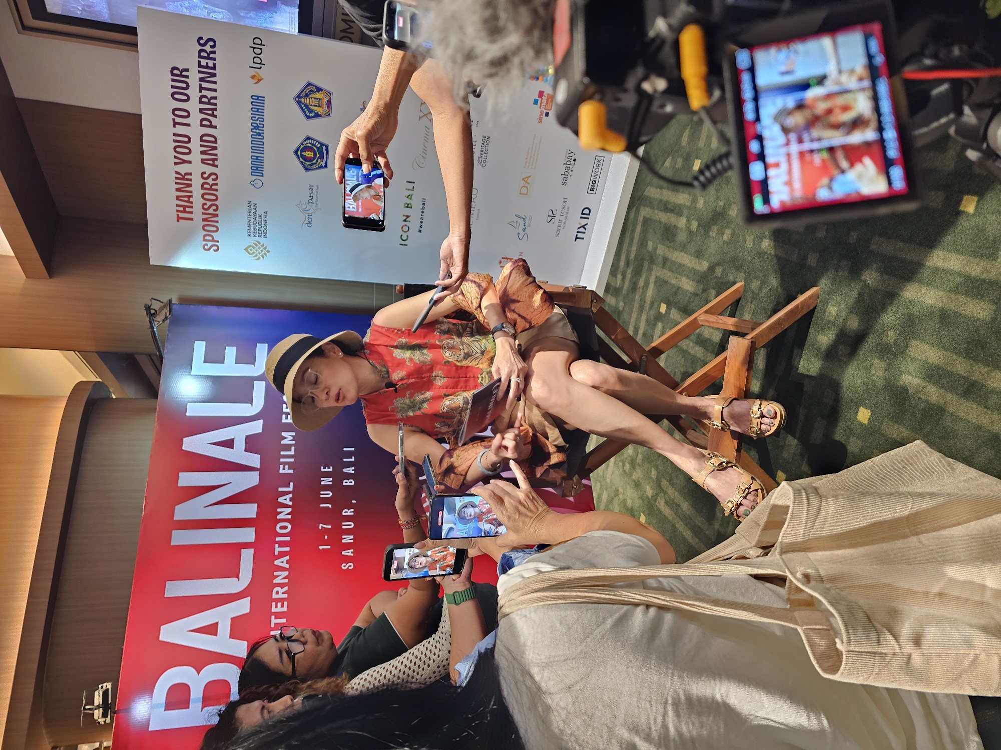 This media file captures an indoor event, likely a press conference or interview, centered around a seated woman being filmed and photographed. The setting appears to be a promotional area with large banners.
The main subject is a woman with fair skin, wearing a wide-brimmed straw hat and a red, short-sleeved collared shirt with a tiger print. She also wears shorts or a skirt, and light-colored sandals with ornate buckles. She is seated in a wooden director's chair, with a book or document on her lap, and appears to be attentive or posing. Her left leg is crossed over her right, revealing her sandals.
Surrounding her are several individuals actively recording the scene. At least four smartphones are visible, held by different people, pointing towards the seated woman to capture video or photos. One phone is held by a hand in the foreground, another by a hand further back, and two more are seen closer to the woman. A professional camera setup with an attached microphone, likely for video recording, is visible in the upper right corner, held by a person whose arm is also visible. The floor is covered with a patterned green carpet.
Prominently displayed behind the subjects are two large upright banners. The primary banner on the left, in red and white, announces "BALINALE INTERNATIONAL FILM F 1-7 JUNE SANUR, BALI," indicating the event is the Balinale International Film Festival taking place in Sanur, Bali, from June 1st to 7th. The second banner on the right, predominantly white with a red top band, reads "THANK YOU TO OUR SPONSORS AND PARTNERS" and lists various logos and names including KEMENTERIAN KEBUDAYAAN REPUBLIK INDONESIA, DANA INDONESIANA, LPDP, den pasar, ICON BALI, #wearebali, Cinema XXI, MERU, The HERITAGE COLLECTION, Sand, DA, SR sanur resort, sababay, TIX ID, and BIGWORK, among others.
The event is taking place indoors, likely during the daytime. The atmosphere appears focused, with participants engaged in documenting the central figure. Given the context of Denpasar, Indonesia, this event is located within Bali.