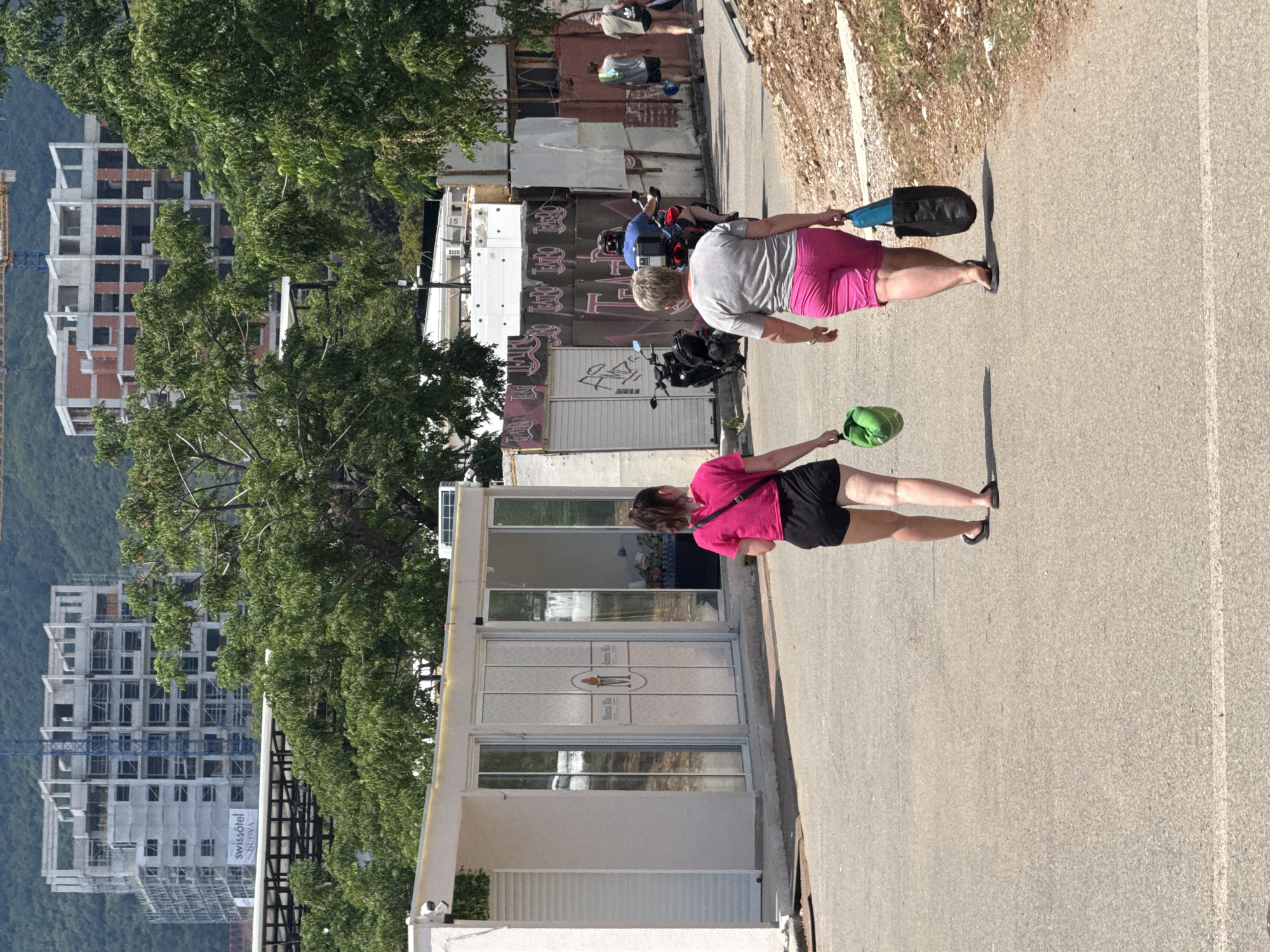 Two women walk away from the camera on an asphalt path in Boreti, Montenegro. The woman on the left wears a bright pink t-shirt and black shorts. She carries a green bag in her right hand. The woman on the right wears a grey t-shirt and bright pink shorts. She carries a blue bag in her right hand and a black bag in her left hand. Both women wear flip-flops. Behind them, a scooter is parked next to a building with graffiti. In the background, a multi-story building under construction with scaffolding is visible, along with trees and a hillside. The day appears sunny with shadows cast on the ground.