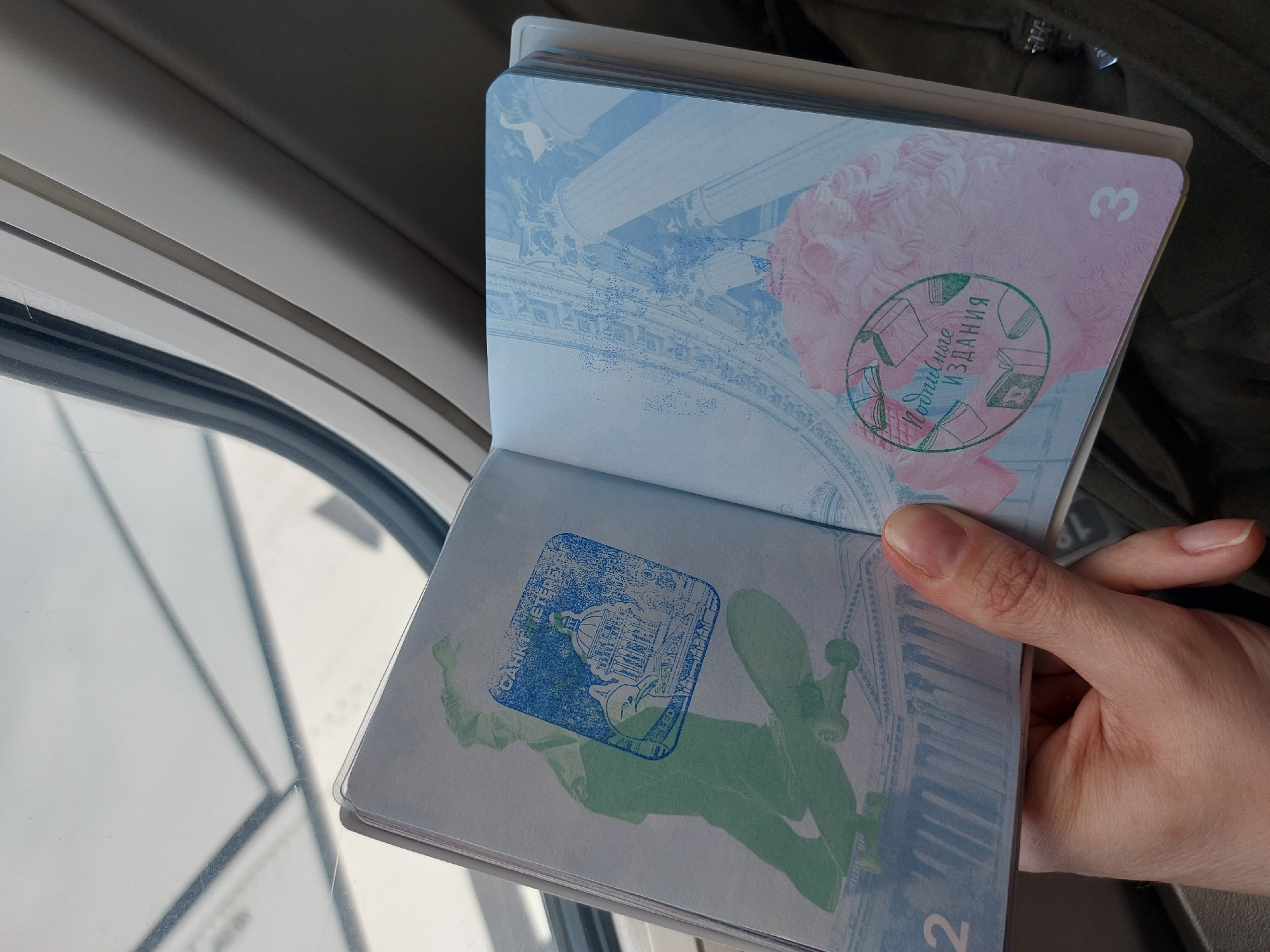A person holds open a passport inside an airplane cabin. The open passport pages display artwork and stamps. One stamp is circular, green, and contains the inscription "Личные издания". The other stamp is square, blue, and includes the text "САНКТ-ПЕТЕРБУРГ", and the image of the Winter Palace and the Alexander Column in Palace Square, located in Saint Petersburg, Russia. The number 2 is on the lower left and the number 3 on the upper right pages. The passport is held against a backdrop of the airplane window and cabin interior.