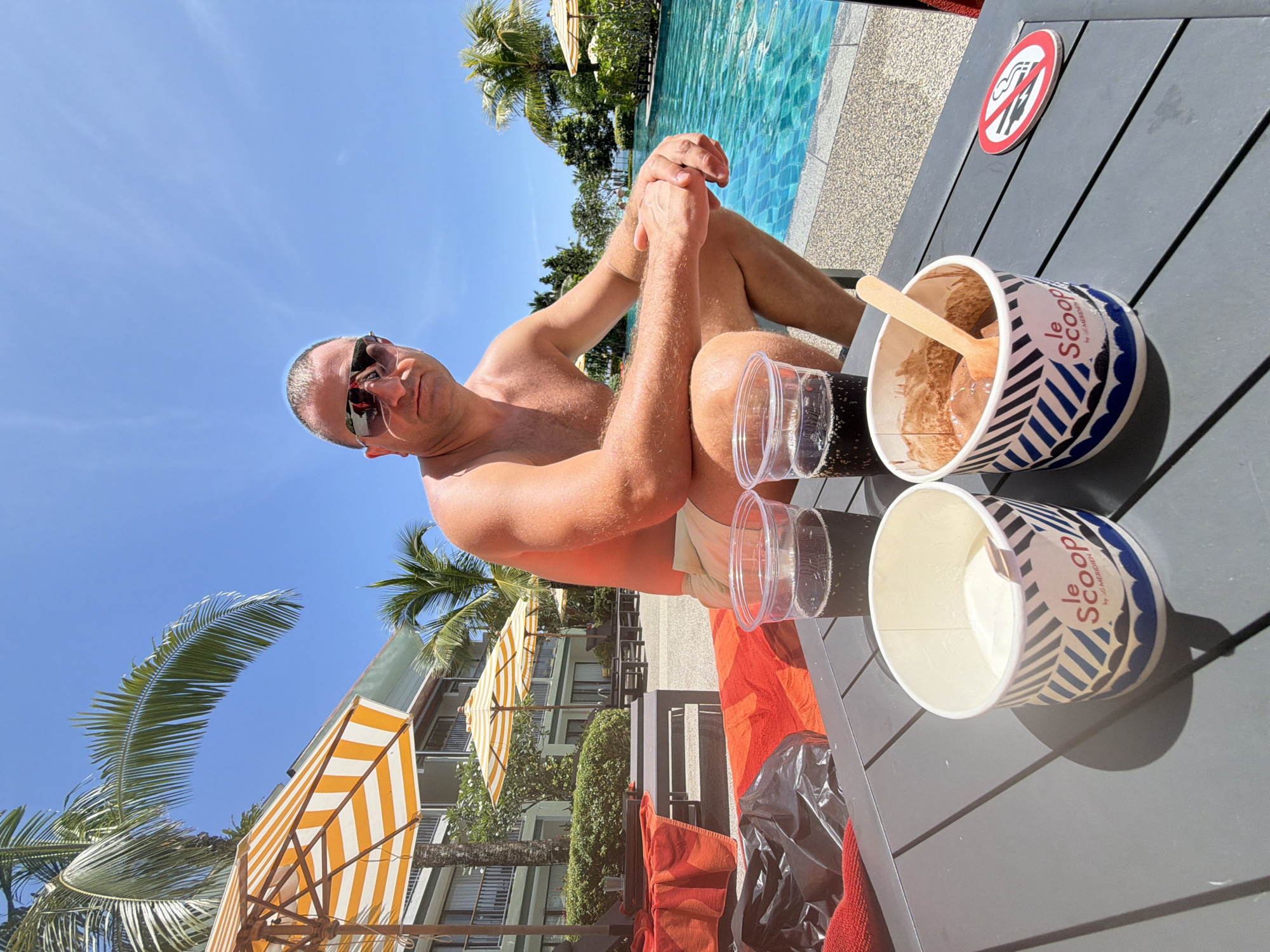 A man is sitting by a pool in Bang Muang, Thailand. He is wearing sunglasses and is shirtless. In front of him on a table are two cups of ice cream, two cups of dark soda, and a trash bag. The pool is to his right, and palm trees and umbrellas are visible in the background. The sky is blue and clear.
