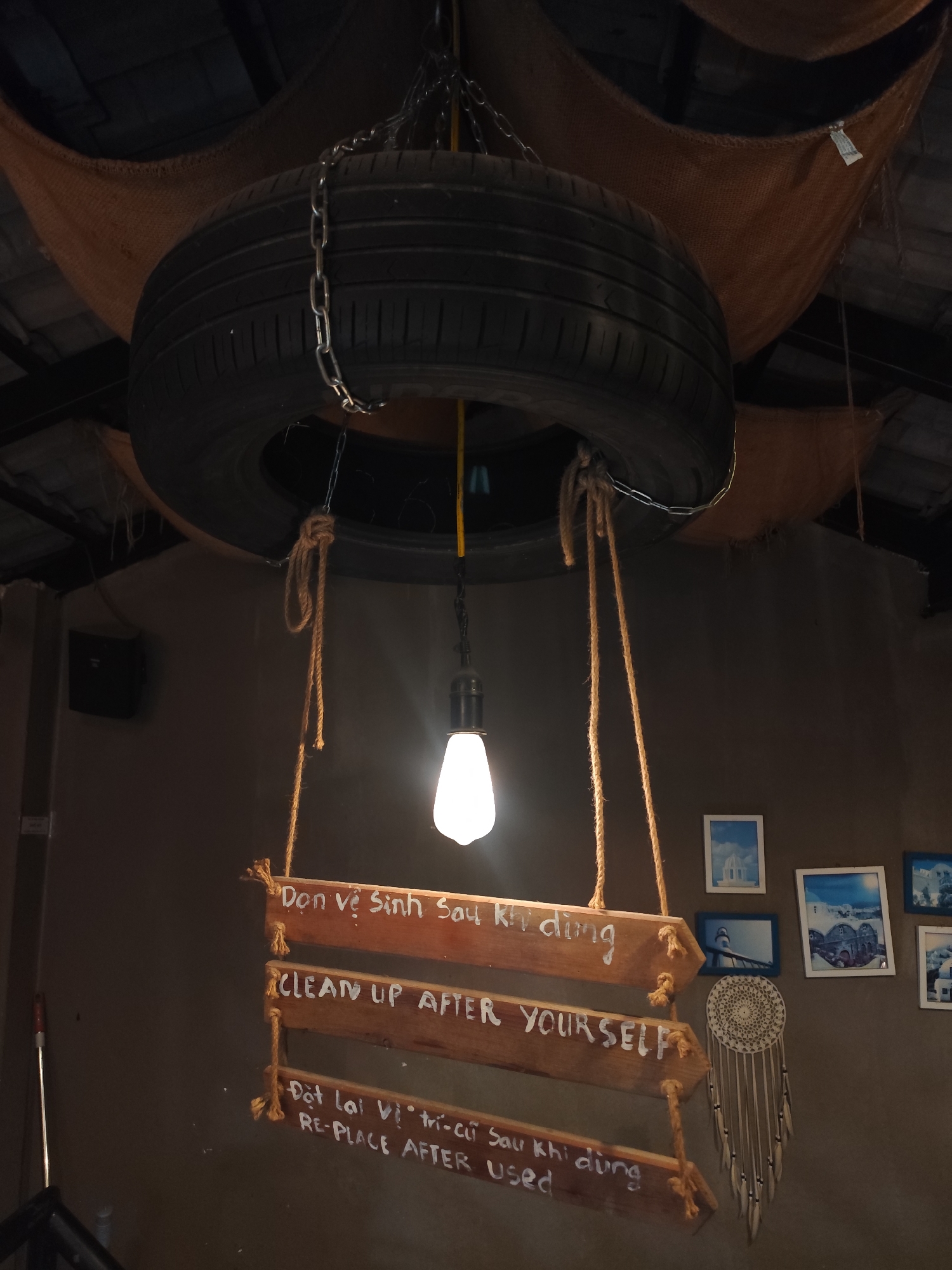 The image depicts a decorative lighting fixture in what appears to be a restaurant or café, possibly in Da Lat, Vietnam. A tire suspended from the ceiling by chains and ropes serves as a decorative element. Hanging beneath the tire is a light bulb, illuminating a series of wooden planks painted with instructions in both Vietnamese and English: "Dọn vệ sinh Sau khi dùng / CLEAN UP AFTER YOURSELF" and "Đặt lại vị trí cũ Sau khi dùng / RE-PLACE AFTER used." These planks are also suspended by rope. To the right of the wooden sign, a wall is decorated with framed pictures and a dreamcatcher. The overall aesthetic suggests a rustic, repurposed design.