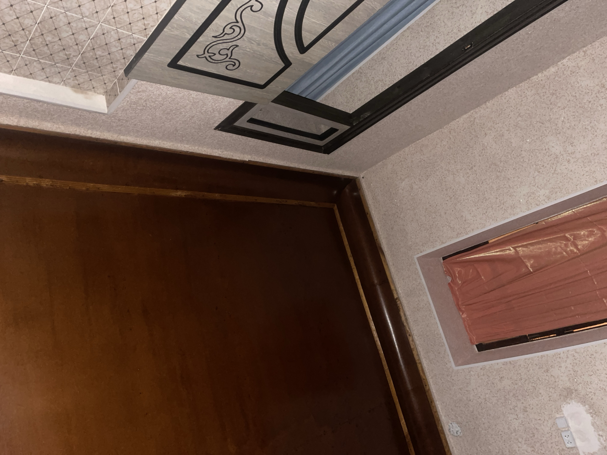 The image depicts an interior scene, likely a hallway or entryway, with a view looking upwards. The dominant features are a large, dark brown wooden panel that occupies the upper left portion of the frame, possibly a ceiling or a large cabinet. Below this, a textured, light-colored wall with a speckled pattern extends across the scene.
To the right of the wooden panel, there is a doorway or a decorative niche. This opening is outlined with a lighter colored trim and contains a vibrant, rust-colored curtain, made of a shiny, plastic-like material. The curtain appears to be covering the entrance to a room or closet.
Further to the right, another decorative element is visible on the wall, featuring a geometric pattern with black outlines and a gray, marble-like inlay. A portion of what appears to be a door or a decorative panel with an ornate scroll design is also visible in this area.
In the lower left corner of the image, there are two white electrical outlets on the textured wall, one of which has a small, white patch of paint or plaster nearby. There is also a circular white object attached to the wall, possibly a smoke detector or a light fixture.
The overall lighting suggests an indoor setting, with light sources not directly visible but contributing to a somewhat dim ambiance. The perspective is from a low angle, looking upwards towards the ceiling and the top portions of the walls and doorway. The specific location within Mangyt, Kyrgyzstan, cannot be determined from the visual information alone.
