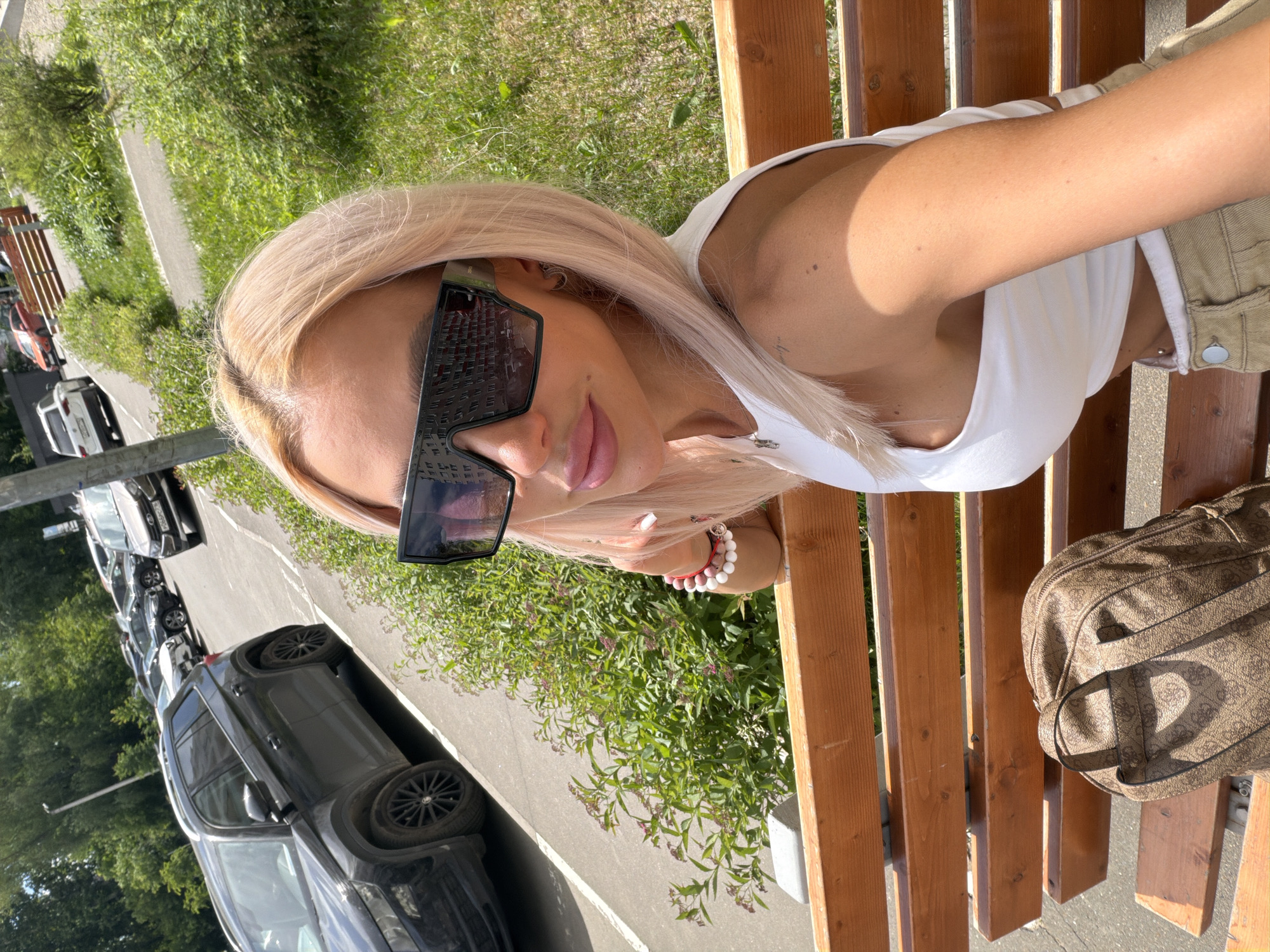 This image captures a fair-skinned female individual seated on a wooden slatted bench under direct sunlight in Kazan, Republic of Tatarstan, Russia. The location is identified by the "116" regional code visible on the license plate "X066PC 116" of a light brown sedan parked on the street.
The woman has long, straight, light blonde hair and is wearing oversized, dark, rectangular sunglasses that reflect a multi-story building. Her lips are full and appear to have a light pink gloss. She wears small hoop earrings. Her attire consists of a white sleeveless crop top and light brown/khaki pants. On her left wrist, she wears two bracelets, one composed of red and white beads, and another of entirely white beads. Her fingernails are painted white. A small, indistinct tattoo is visible on her left shoulder/upper arm.
To her right, on the wooden bench, rests a brown backpack patterned with the "GUESS" logo.
The background features a paved asphalt road lined with parked vehicles. Immediately to the left of the woman, a black Skoda Kodiaq SUV is prominent. Further down the street, various other cars are parked, including a white SUV and the light brown sedan with the identifiable Russian license plate. Beyond the road, green bushes and trees are visible, bordering what appears to be a park or green space. A metal railing and a second wooden railing define the boundaries between the road and the green areas. A street light pole stands on the left side of the road. The bright lighting and pronounced shadows indicate a sunny day.