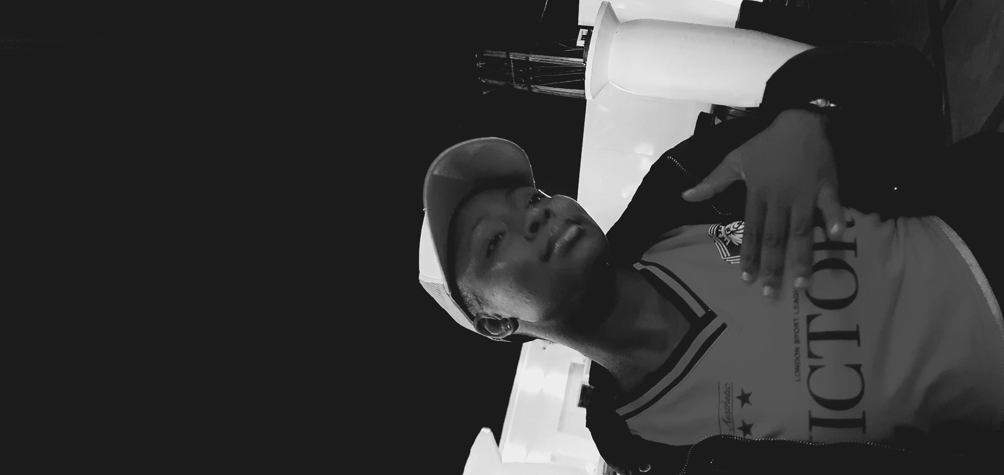 In a black and white photograph, a person wearing a baseball cap and a t-shirt with "VICTOR" printed on it is shown from the chest up. The person is looking towards the camera with a slight smile and their right hand is raised, with fingers splayed, resting on their chest. The background is dark, with some blurred white structures and poles visible. The image is taken at night. The location is not discernible from the image.