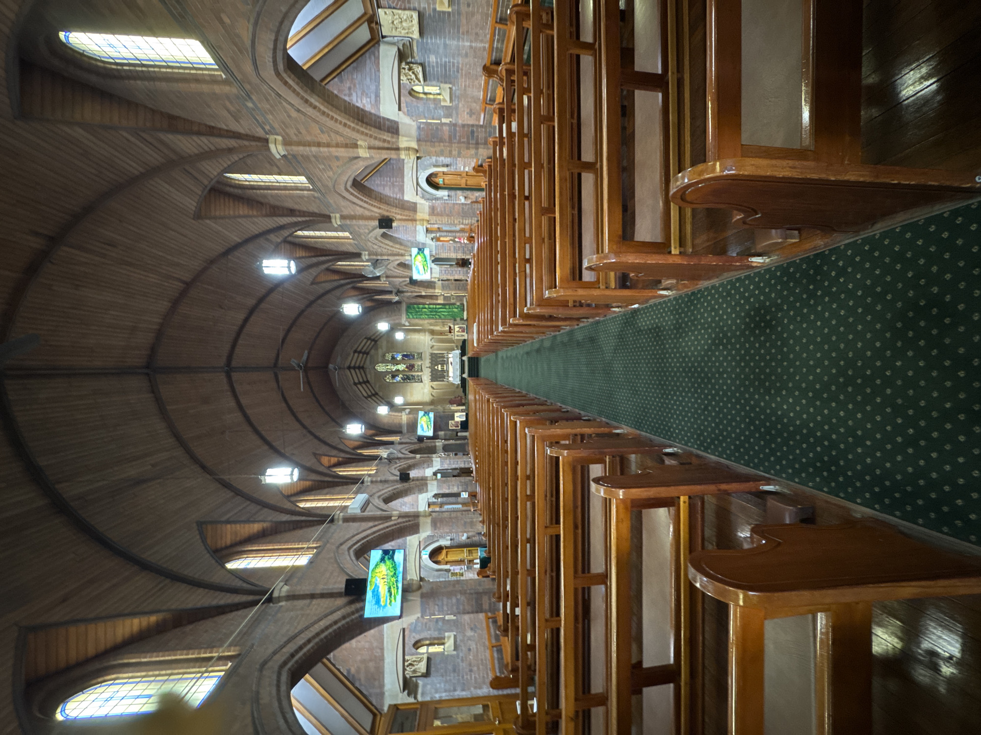 The image shows the interior of St. Michael's Anglican Church in Vaucluse, Australia. The perspective is from the back of the church, looking towards the altar at the front. Rows of polished wooden pews fill the nave, separated by a dark green carpeted aisle. The ceiling is a vaulted, timber construction. Brick arches and columns line the walls, with stained-glass windows visible in the upper sections. Two large digital screens are mounted on the left wall, displaying what appears to be animated images. No people are visible in the scene.