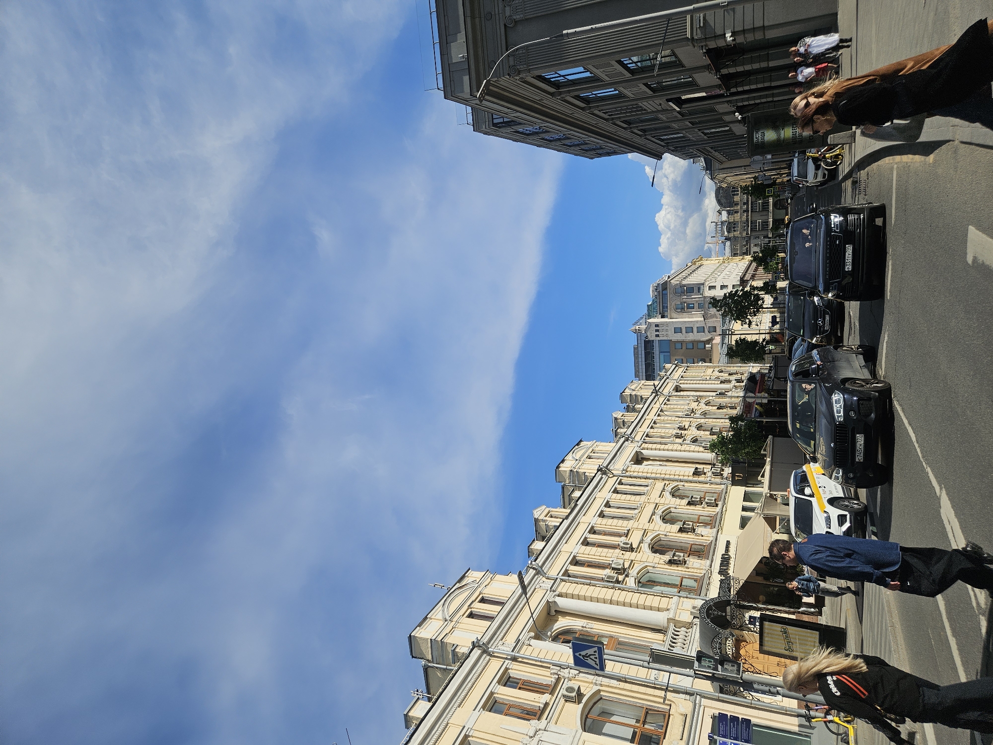 The image depicts a street scene in Moscow, Russia, with buildings lining both sides of the street. The sky is blue with scattered clouds. Traffic consists of cars and a taxi, with pedestrians walking along the sidewalks. The buildings are a mix of older and more modern architecture, some with ornate facades. The overall atmosphere suggests a busy urban environment on a sunny day.