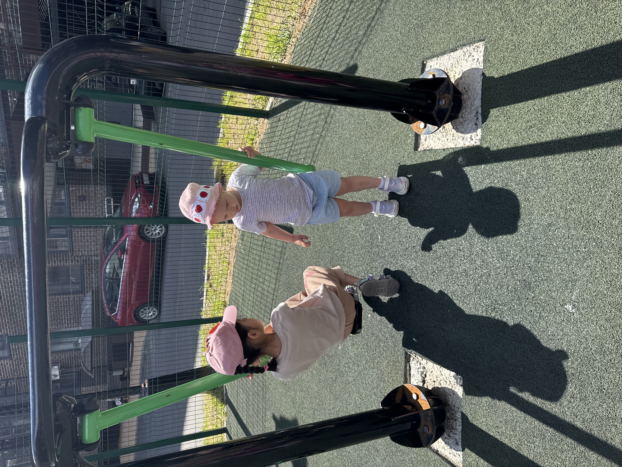 The image captures two young girls on a playground in the city of Vyazniki, Russia, on a sunny day. One girl, wearing a pink baseball cap and a beige t-shirt with beige pants, is seated on a swing and appears to be in motion. The other girl, sporting a pink hat adorned with hearts and a striped t-shirt with light blue shorts, stands beside the swing, holding one of the green support poles. Both girls are wearing light-colored sneakers. The playground features a rubberized green surface and is enclosed by a dark metal fence. In the background, a red car is parked on a street adjacent to a brick building. The shadows of the girls and the swing set are cast on the playground surface, indicating a bright sunlit environment.