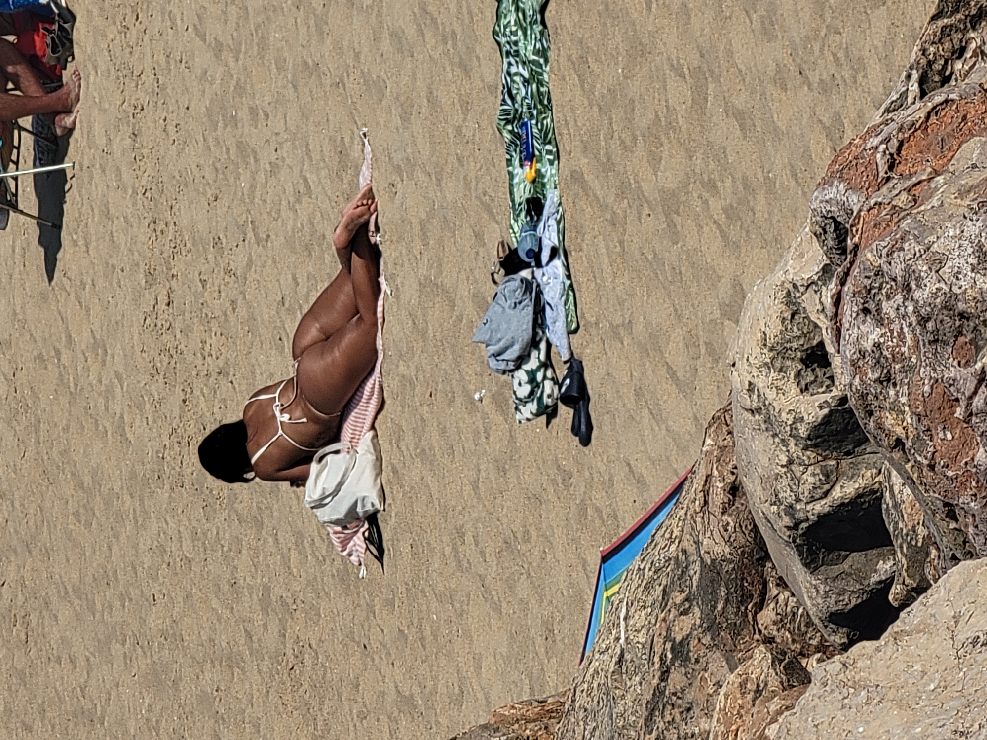 The image shows an overhead view of a sandy beach. A person is lying on their stomach on a towel, facing away from the camera. They are wearing a white bikini. Next to the person, a white bag and a striped towel are visible. Further up the beach, several pieces of clothing are hanging from a rope or line, including a green and white patterned fabric, a light blue shirt, and dark shorts. The beach is textured with ripples from the wind. To the right of the image, a rocky outcrop juts into the scene, with rough, textured surfaces in shades of brown, grey, and white. Part of a colorful umbrella or beach shelter with red, blue, and yellow sections is visible at the base of the rocks. The lighting suggests it is daytime, likely sunny, with shadows cast on the sand. The scene depicts a relaxed day at the beach. No text is visible in the image.