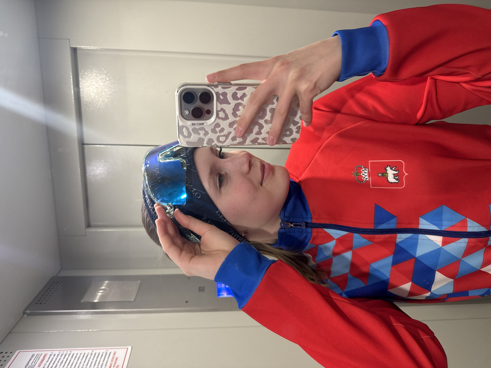 A person in a red jacket with a blue and white geometric design and blue cuffs is taking a selfie in what appears to be an elevator. The person is wearing ski goggles on their head and is holding a smartphone with a white and gray leopard-print case. The jacket has a crest or emblem with a crown and a white animal, possibly a bear, on the right side. The elevator interior has gray or white walls and a metal panel. A sign with text in a foreign language is visible on the lower left side of the image. The location is possibly in Chaikovsky, Russia.