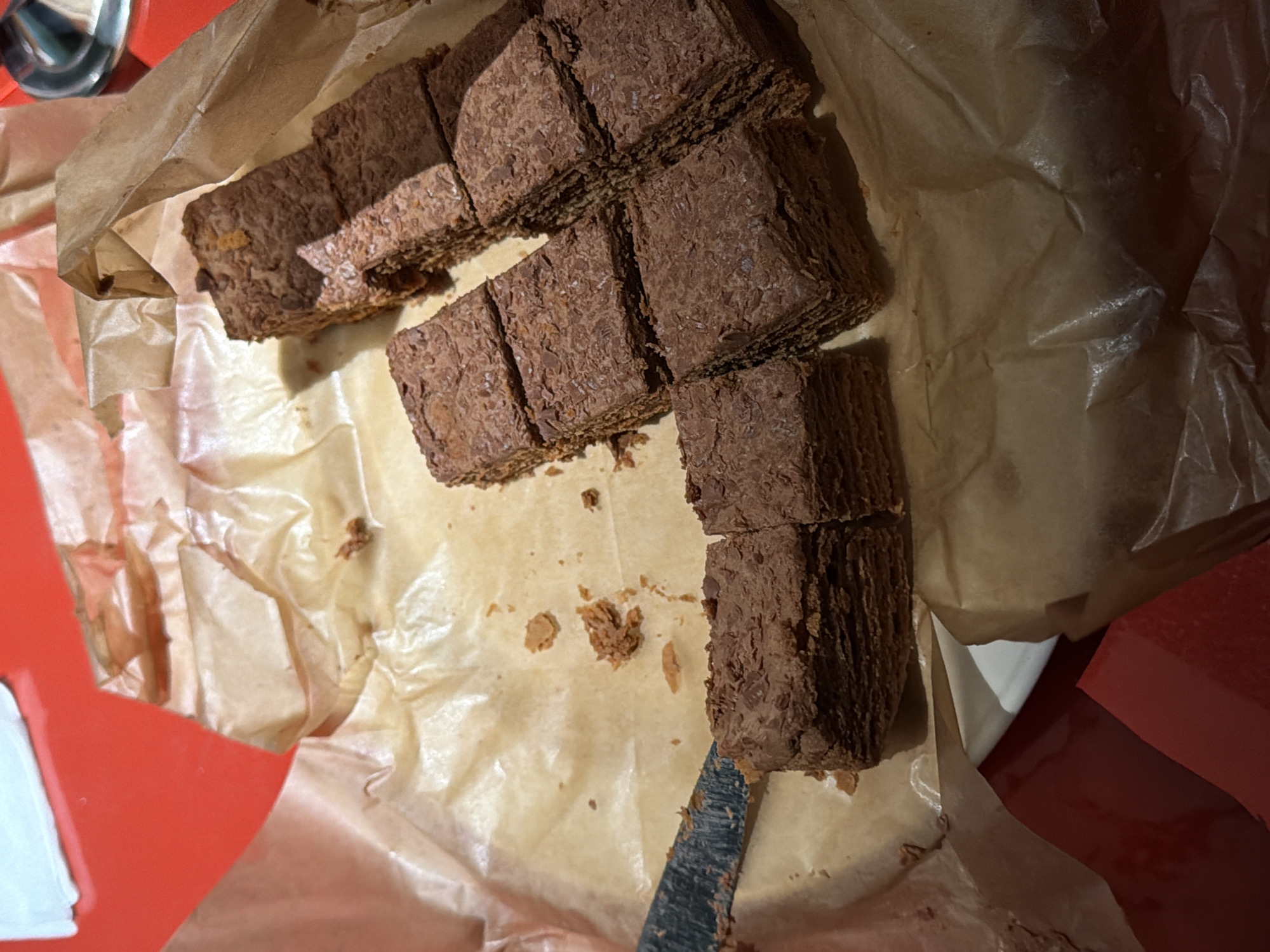 The image depicts a collection of layered desserts, likely brownies or similar baked goods, arranged on a sheet of parchment paper. The desserts are rectangular and appear to have a textured surface. A knife is present near the desserts, suggesting they were recently cut. The parchment paper is crumpled and stained, possibly with residue from the desserts. The background consists of a red surface and white paper. The image does not provide any information to determine the location of the photograph in Yerevan, Armenia.