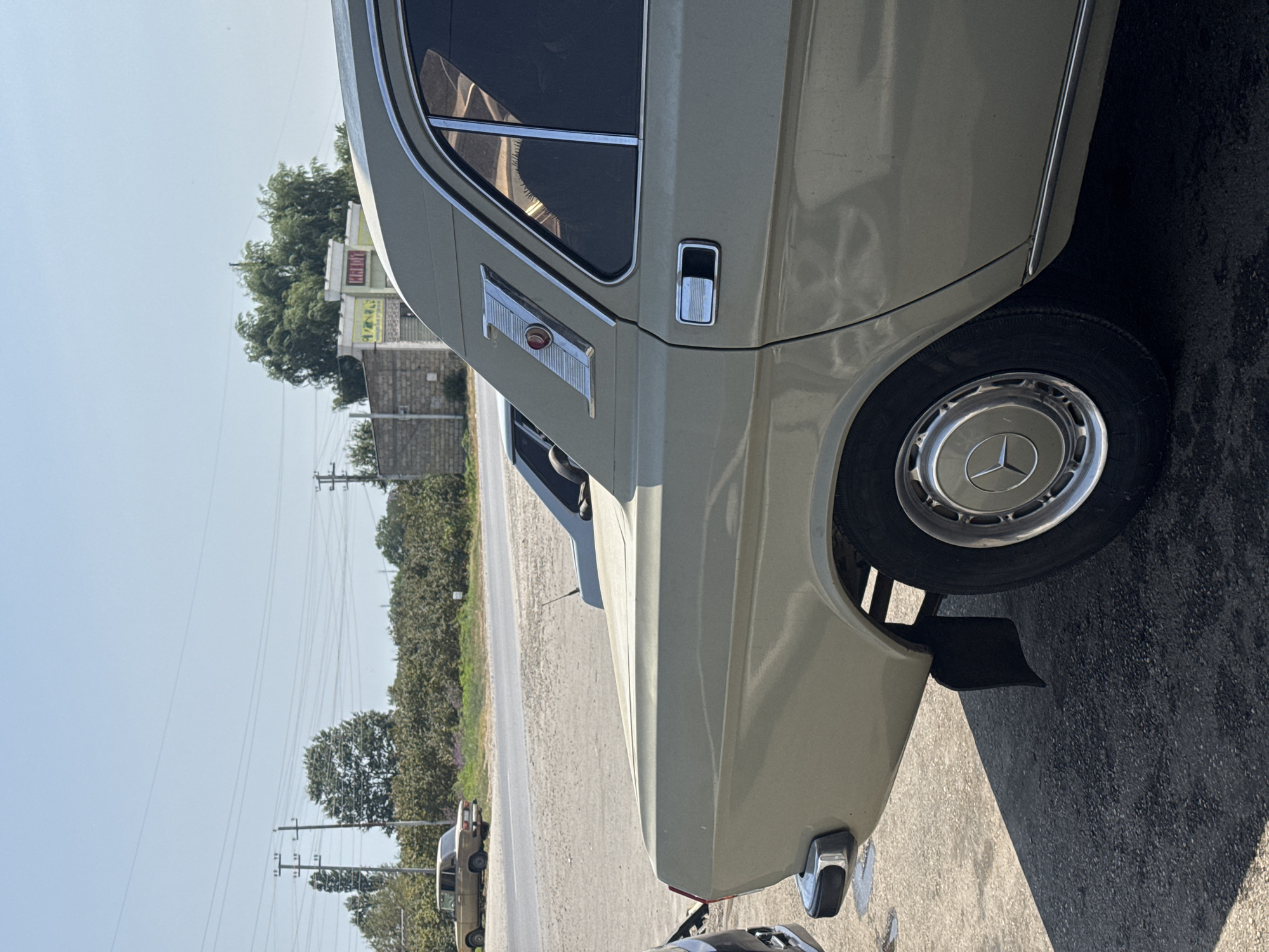 This is a medium-angle, close-up, eye-level, full-color, outdoor photograph of the rear passenger side of a tan sedan parked on a paved surface. The car has a chrome trim accent above the rear wheel arch with a red circular reflector and a stylized "Z" emblem. The rear bumper is chrome with black end caps. A mud flap is visible behind the rear wheel, which has a silver hubcap with a circular emblem. The car's windows are tinted dark. Another light blue car is partially visible in the background, to the left of the tan car, with its rear wheel also visible. In the distance, a paved road runs parallel to a line of trees and bushes. Across the road, there are power lines supported by utility poles. Behind the trees, a building with a light-colored facade and a yellow sign with red lettering reading "MƏLDİ" is visible. The sky is clear and bright blue. The scene is set in the city of Müşkür, Azerbaijan.