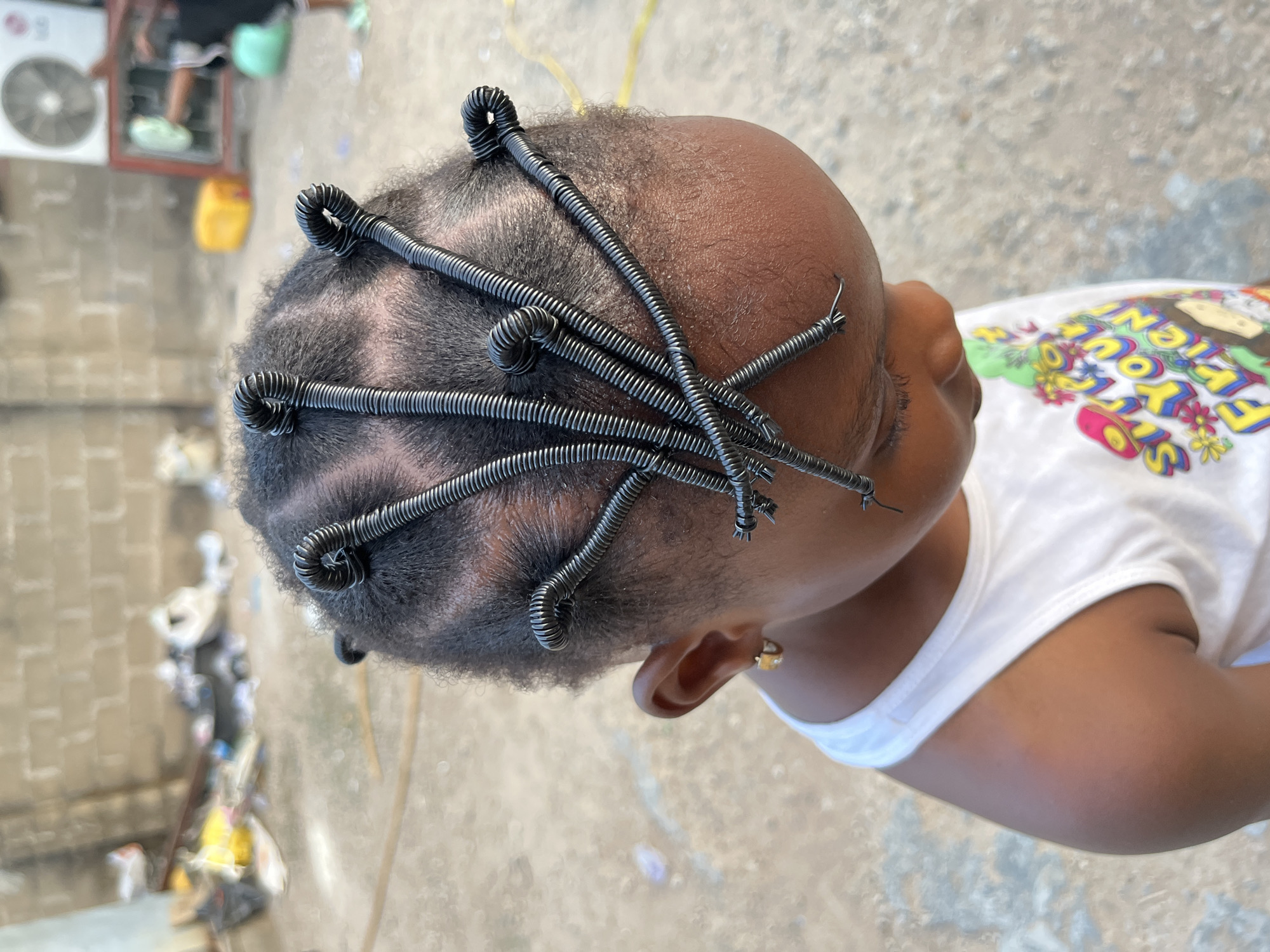 The image shows the top of a child's head with a hairstyle of cornrows and metal coils wound around the braids. The child wears a white t-shirt with cartoon graphics. The background includes a concrete or paved area, possibly a street or yard, with some trash and debris visible. In the distant background, there's part of a building with an air conditioning unit. Given the prompt specifies the location as Madalla, Nigeria, the setting is an outdoor location typical of a residential or commercial area in that city.