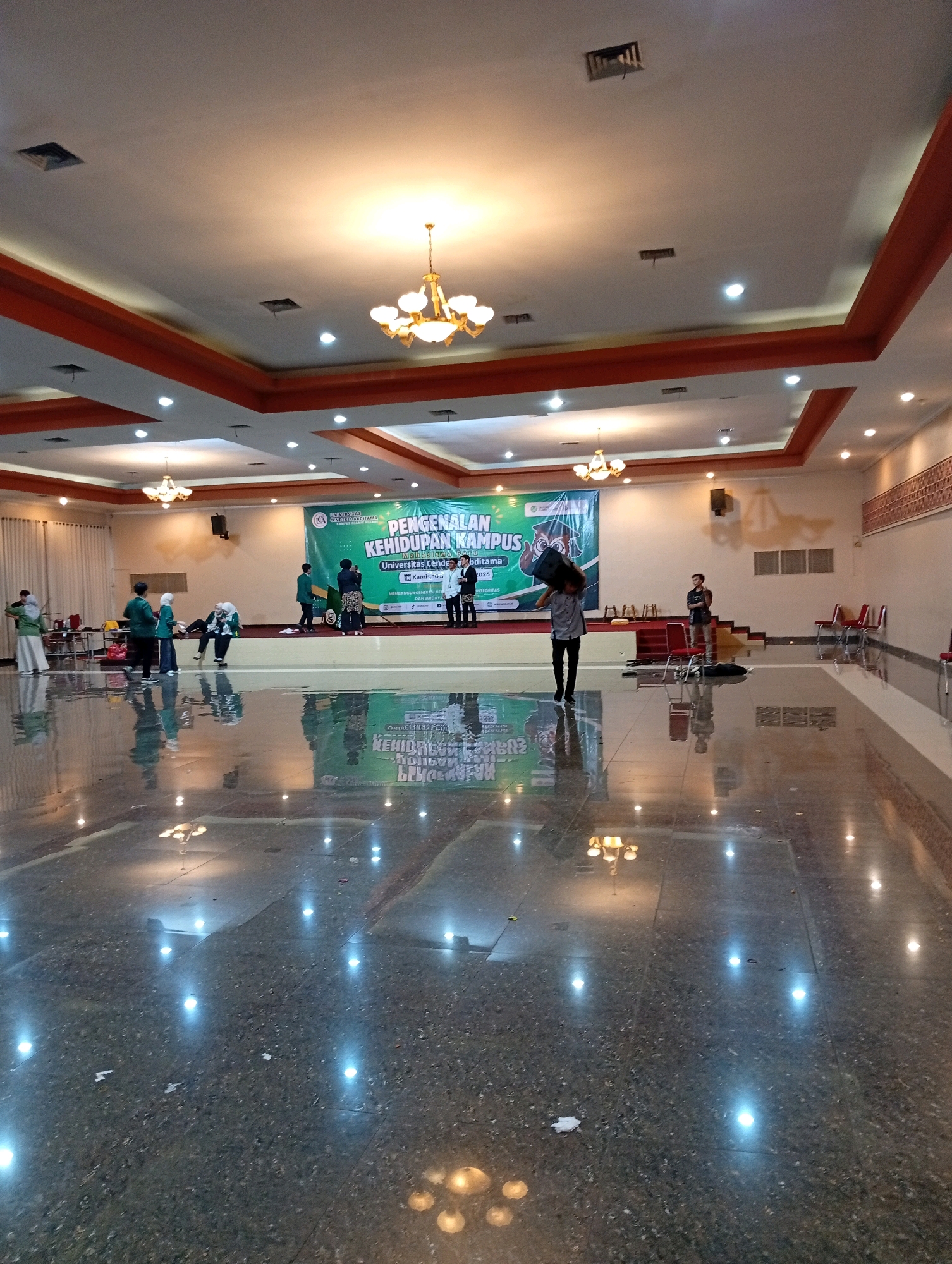 The image depicts an indoor event space with a polished, reflective floor. In the background, a stage is set up with a large green banner that reads "PENGENALAN KEHIDUPAN KAMPUS" (Introduction to Campus Life). Several people are visible on and around the stage, some appearing to be participants or staff. To the left of the stage, a group of individuals in casual attire are gathered. To the right, there are rows of red chairs and a person standing on the stage. The ceiling features recessed lighting and decorative chandeliers. The overall impression is of a venue prepared for an academic or organizational event. The location is identified as Kelapa Dua, Indonesia.