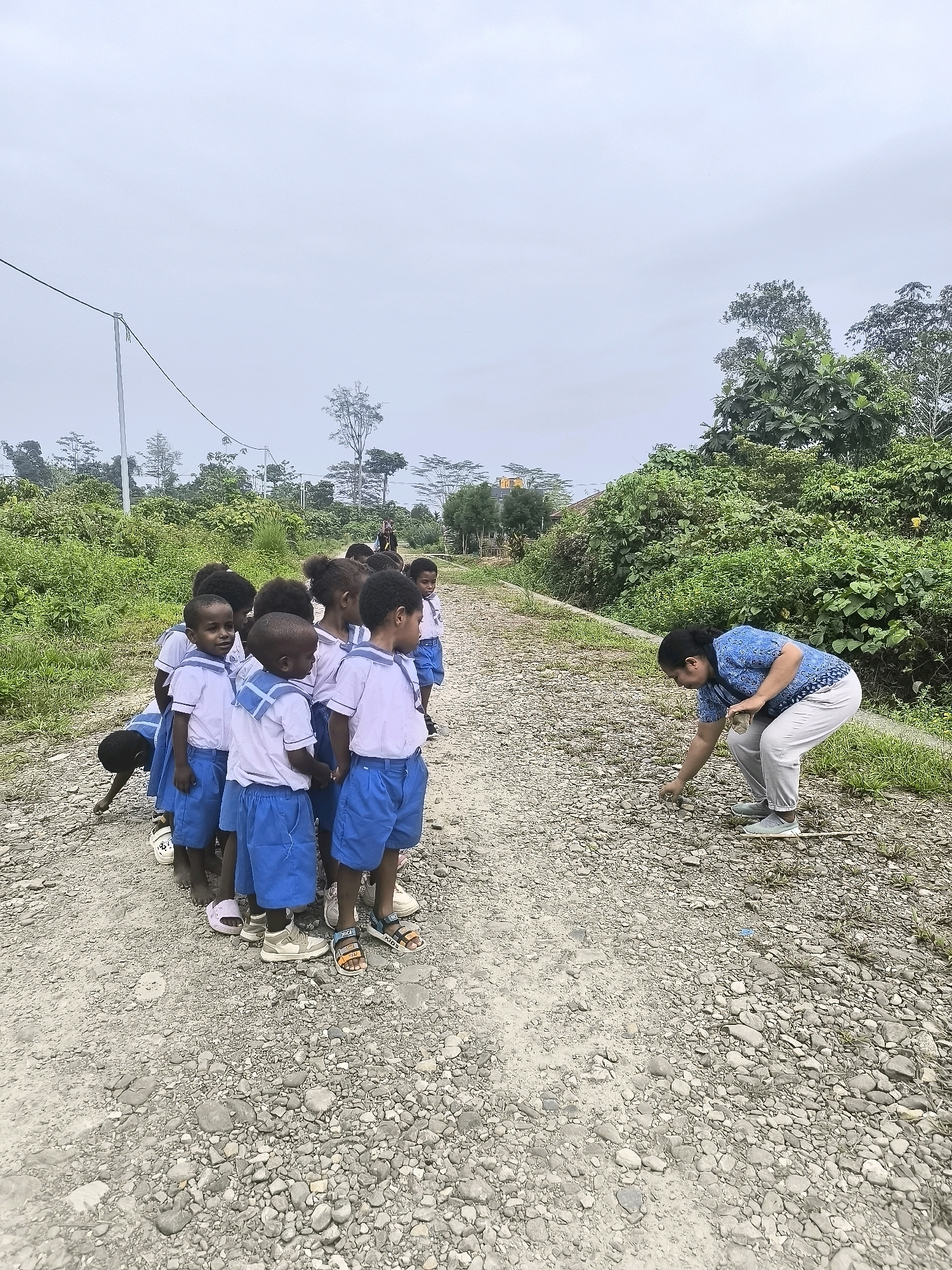 In the city of Timika, Indonesia, on a cloudy day, a group of schoolchildren in blue and white uniforms stand in a line on a dirt road, facing a woman in a blue shirt and light-colored pants. The woman is bending down, picking up small stones from the road. The children, all of African descent and appearing to be between the ages of 5 and 8, are looking in various directions, some towards the woman, others off to the side. One child is partially lying down in the background. The road is made of gravel and dirt, with green vegetation lining its sides and extending into the background, where trees and a few houses are visible. Power lines are strung overhead. The overall scene suggests an outdoor educational activity or a break during a school day in a rural or semi-rural setting within Timika.