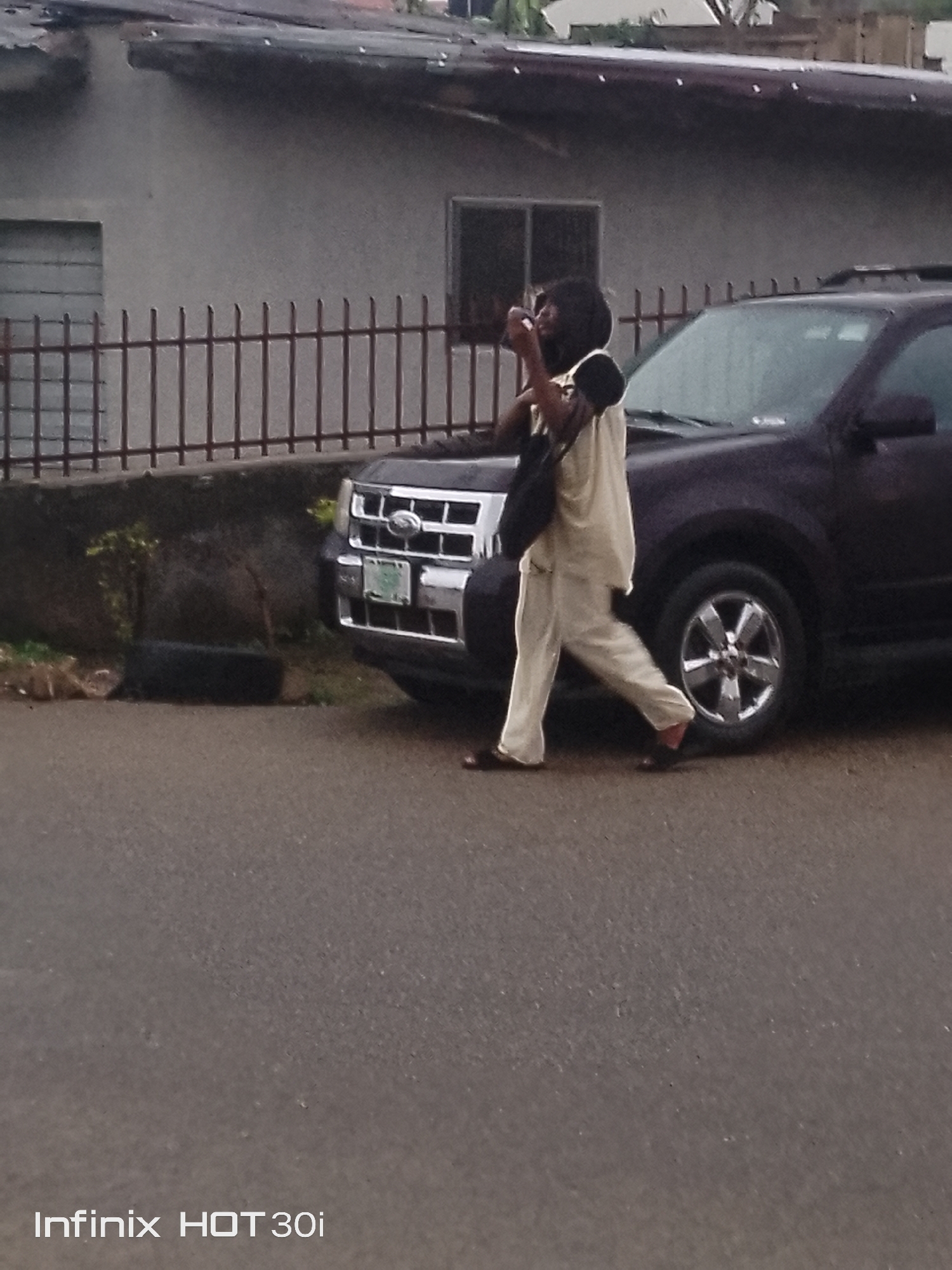 An individual wearing a light-colored outfit walks in front of a dark-colored SUV parked on a paved road. The person is holding a dark bag over their shoulder and appears to be looking at a device in their hand. In the background, a light-colored building with a dark roof is visible behind a metal fence. A small plant is located in front of the fence. The scene appears to be in a residential or urban setting.