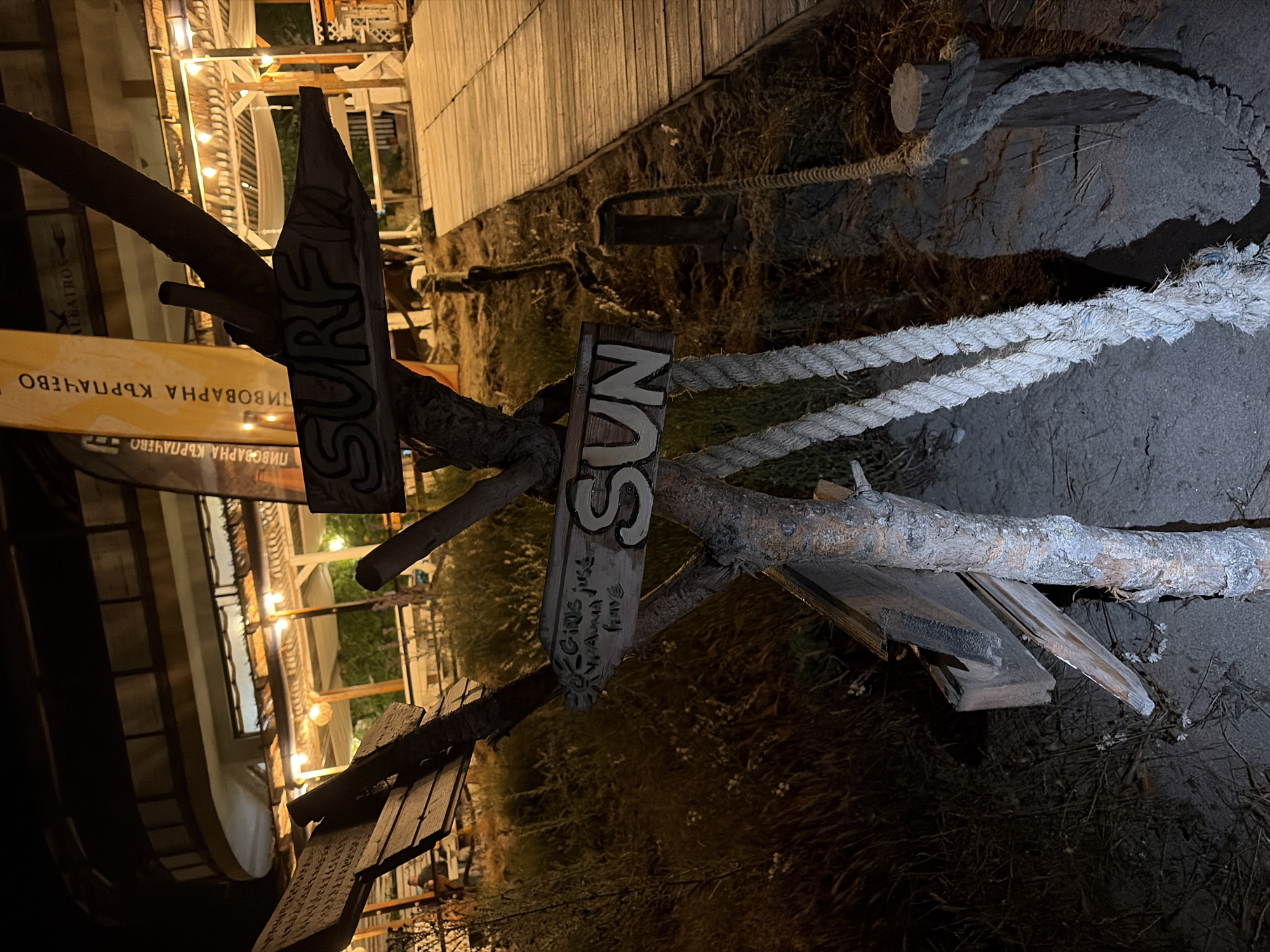 The image depicts a nighttime scene at a beachside establishment in Tsarevo, Bulgaria. In the foreground, a rustic wooden signpost stands on sandy ground interspersed with dry grass. Attached to the post are three wooden arrow-shaped signs. The upper right sign points right and reads "SURF" in white letters. Below it, another sign points right and reads "SUN" in white letters. To the left, a sign reads "GIRLS just wanna have fun" with "S" being the start of "SUN" on the same plank, indicating a playful direction towards the sun/beach. Thick, braided ropes are draped around the base of the signpost and extend into the ground, with one segment wrapped around a small wooden bollard next to a wooden boardwalk.
The wooden boardwalk extends from the midground on the right, receding into the distance. To the left of the boardwalk and beyond the signpost, an outdoor dining or lounge area is visible, illuminated by numerous string lights hanging under a covered structure. Several indistinct figures of people are present in this area, with one person clearly visible seated at a table on the far left. In the background, a multi-story building with a distinctive curved facade on its upper level is illuminated. A tall, vertical banner with Cyrillic text, reading "ПИВОВАРНА КЪРПАЧЕВО" (Karpachevo Brewery) and "ALBATROS," is visible to the left of the building, suggesting a commercial establishment like a restaurant or bar. The scene is lit predominantly by artificial warm lights, casting shadows and creating a relaxed evening ambiance.