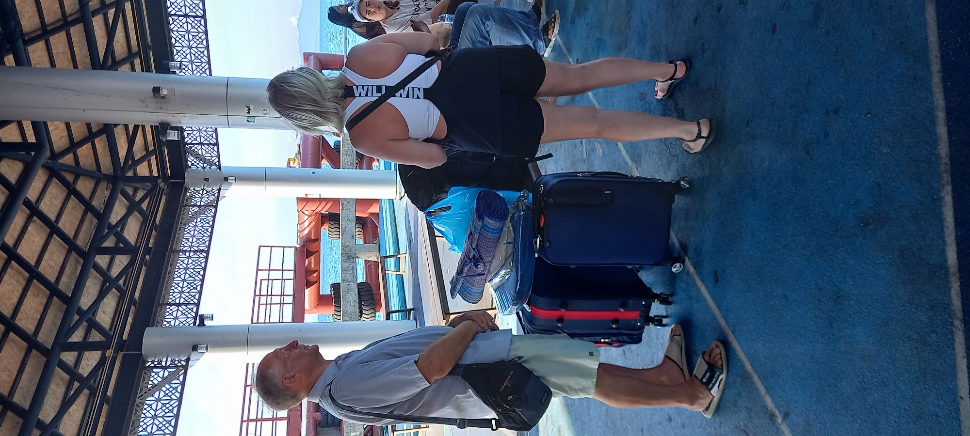The image shows a group of people on a ferry, likely in Thong Sala, Thailand. A man in a light blue shirt and khaki shorts stands next to two pieces of luggage on a blue floor. A woman in a white sports bra with the words "WILL WIN" and black shorts stands nearby. Other passengers are seated in the background. The ferry has metal support beams and a covered roof with patterned edges. A view of the sea is visible between the pillars.