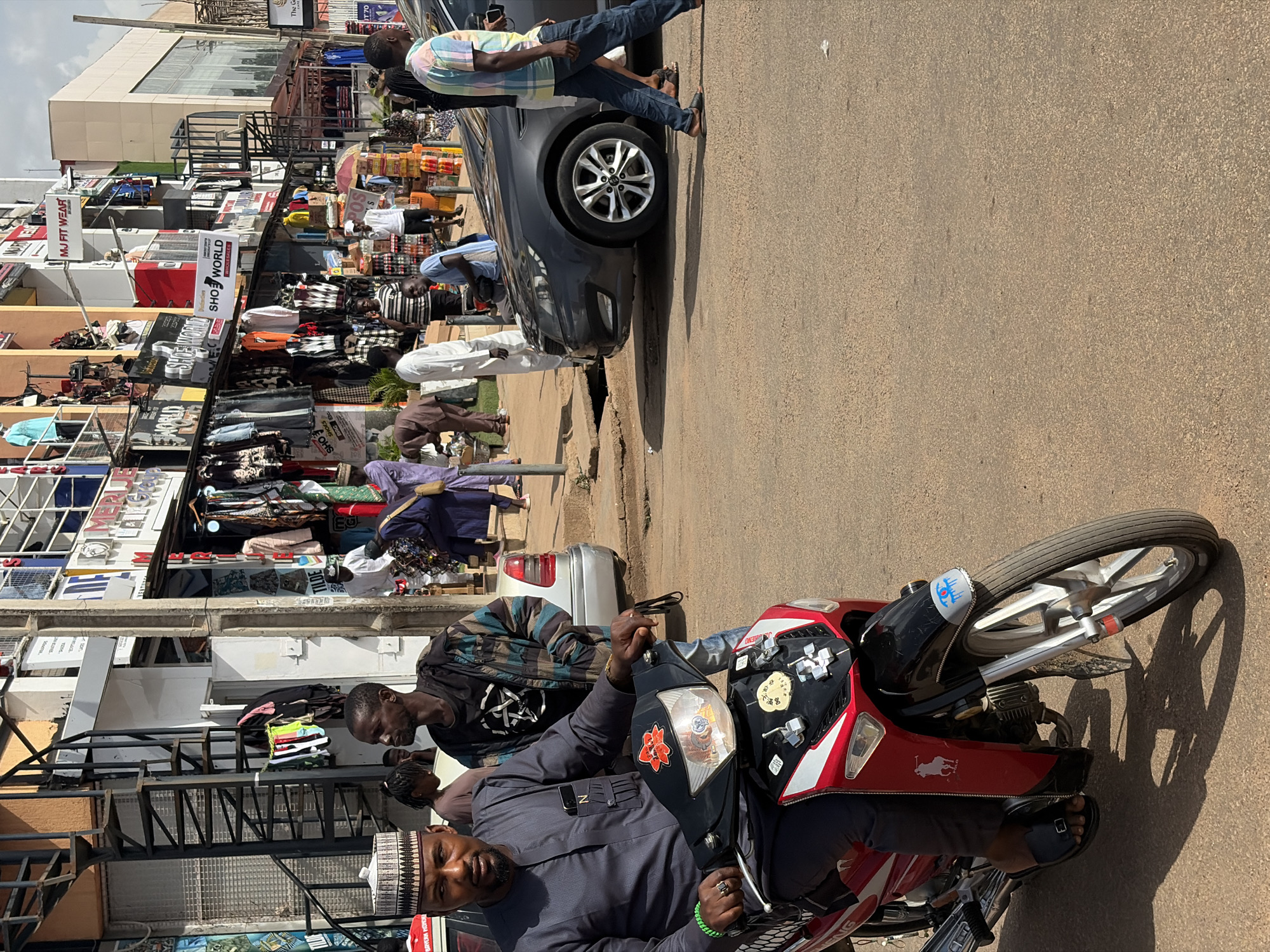 A medium shot captures a busy street scene in Jos, Nigeria, with a man in traditional attire riding a red and black motorcycle in the foreground. He wears a grey kaftan and a patterned cap, and a green bracelet on his left wrist. Behind him, a silver car is parked on the right side of the road, and a dark grey sedan is stopped on the left. The background reveals a row of shops with displays of clothing and other goods. A spiral staircase is visible on the side of a building, and several people are walking on the sidewalk. The street is paved with asphalt, and the sky is overcast.