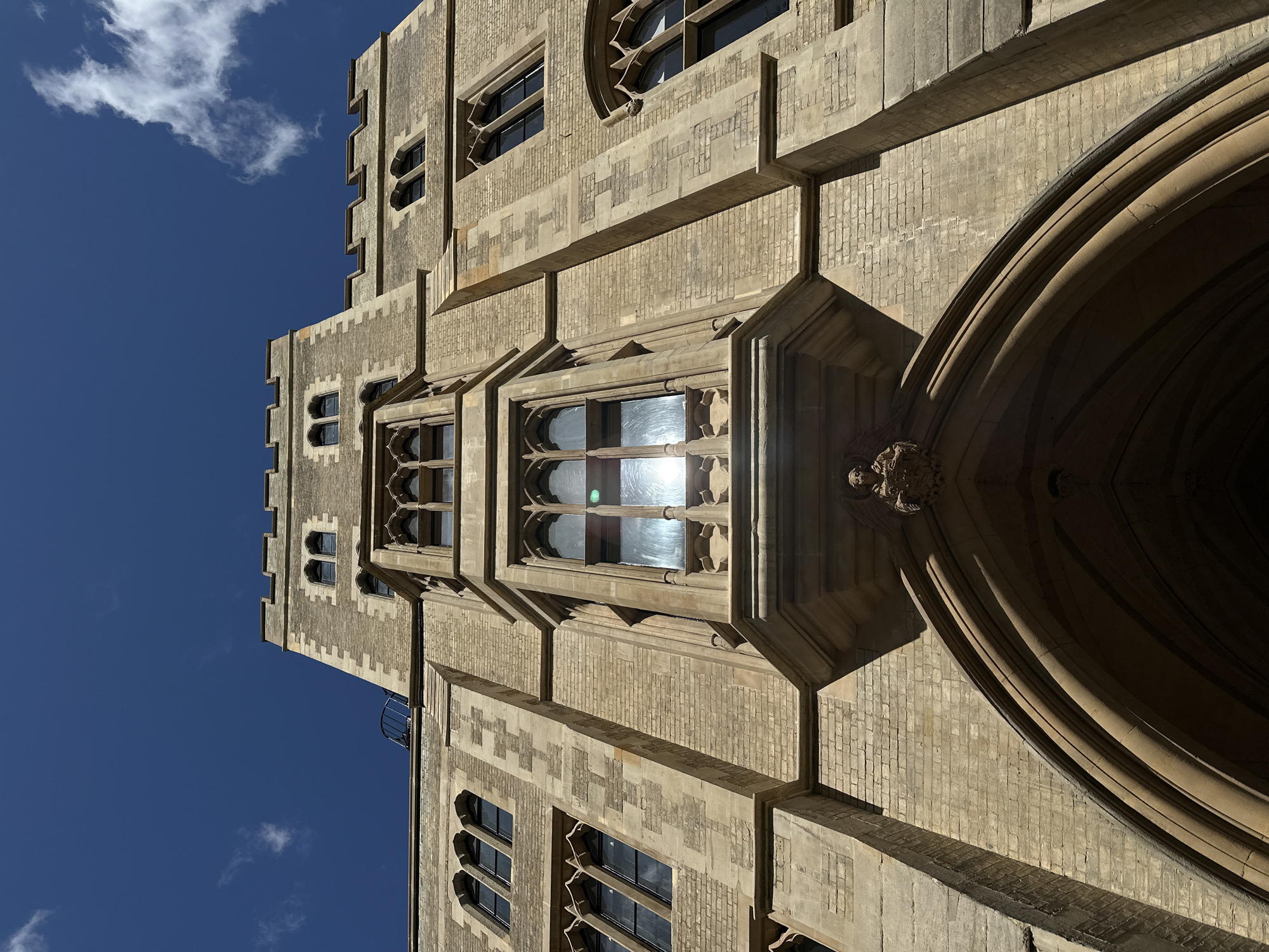 The image shows a low-angle view of a stone building in London, United Kingdom. The building features a tower with multiple windows, and a large arched entrance. The stone facade has a pattern of textured bricks and decorative carvings. The sky is a clear blue with a few scattered clouds. The sun reflects off the windows of the building. There are no people visible in the scene.