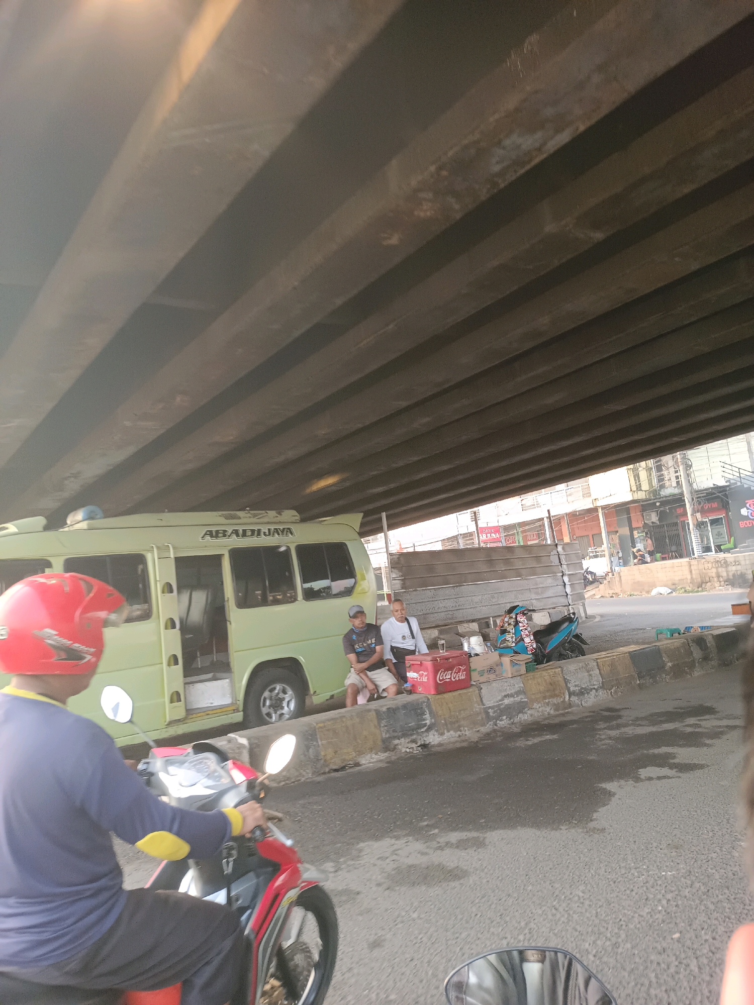The image depicts a street scene in Karawang, Indonesia, under a large overpass constructed of concrete beams. In the foreground, a person wearing a red helmet and a blue long-sleeved shirt is riding a red and black motorcycle. To the left, a light green bus with "ABADJAYA" written on its side is parked. A few people are seated on a concrete barrier next to the bus, some of them near a red Coca-Cola cooler and boxes. A blue and black motorcycle is also parked nearby. The road surface appears wet in some areas, suggesting recent rain or a cleaning activity. The background shows buildings, including one with signage that appears to be for "SPORTS GYM." The lighting indicates it is likely daytime, possibly late afternoon given the angle of the sunlight.