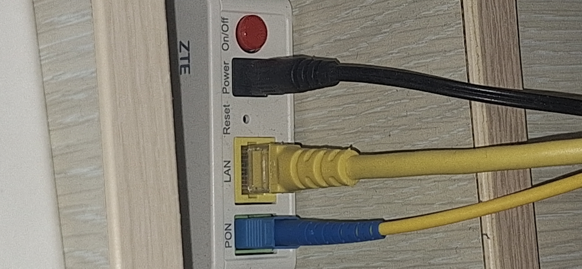 The image shows a close-up of a ZTE network device with several cables connected to it. The device is white and has labels printed on it. From top to bottom, the visible labels are "ZTE", "Reset", "Power", "On/Off", "LAN", and "PON".
There are three cables plugged into the device. The topmost cable is black and connects to a port labeled "Power" or "On/Off". Next, a yellow cable is connected to a port labeled "LAN". This appears to be an Ethernet cable. The bottommost cable is blue and yellow and connects to a port labeled "PON". This is likely an optical fiber cable. A red button, possibly a reset or power button, is visible above the "Power" label.
The network device is mounted on a textured, light-colored surface that resembles wood paneling or a laminate. The overall setting appears to be indoors, likely in a home or office environment in Bila Tserkva, Ukraine, given the provided location context. There are no people visible in the image. The lighting suggests it might be daytime or well-lit interior lighting. The image is focused on the ports and cables of the network device, highlighting the connectivity aspects.