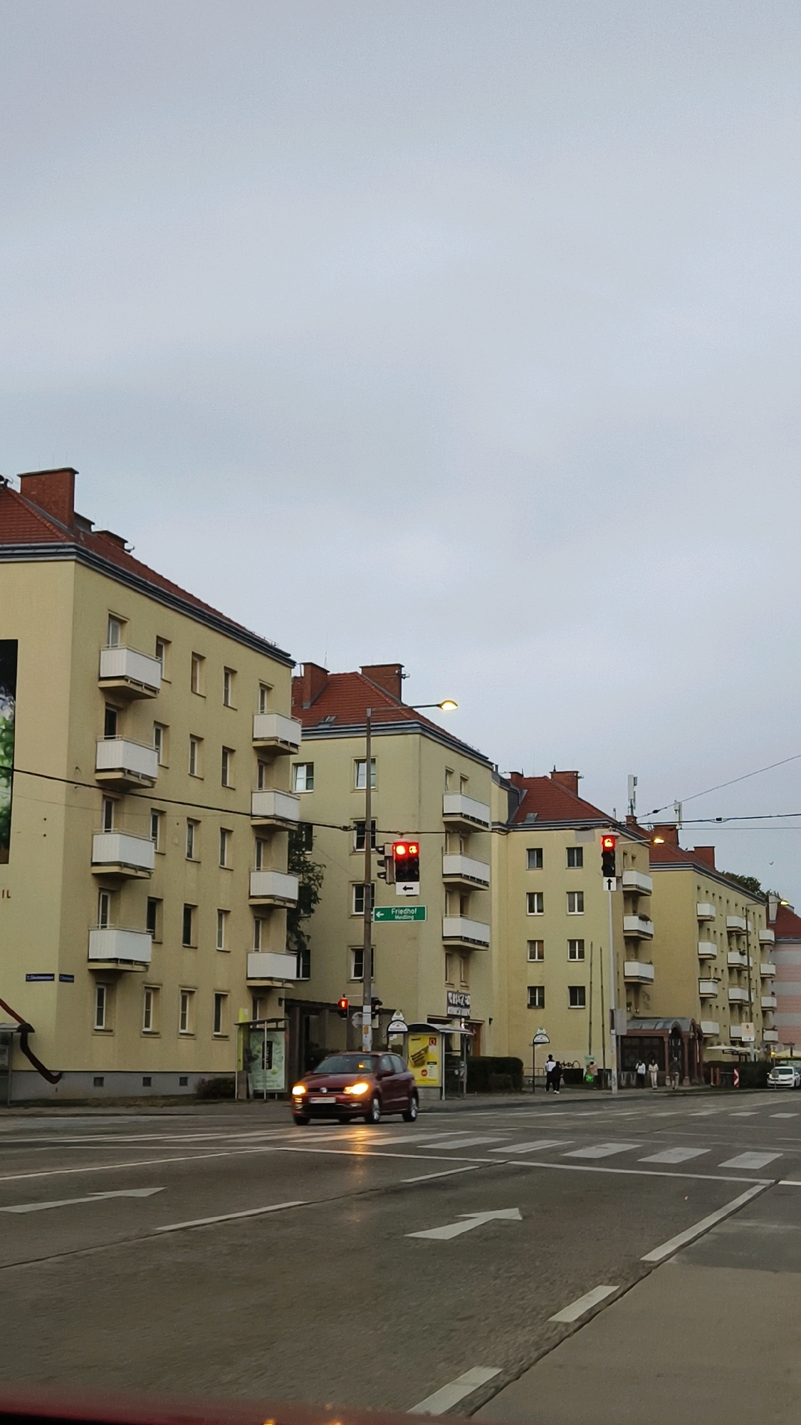 The image captures a street scene in Vienna, Austria, on a cloudy day. A row of pale yellow, multi-story apartment buildings with red tiled roofs lines the right side of the street. White balconies punctuate the facades of these buildings. A red car is stopped at a traffic light on the left side of the road, with pedestrian crossing lines clearly visible in the foreground. Traffic lights are red in both visible directions. A street sign pointing to "Friedhof" (cemetery) is also visible. A street lamp is illuminated overhead. A few pedestrians can be seen near bus stops or building entrances. The overall atmosphere is subdued due to the overcast sky.