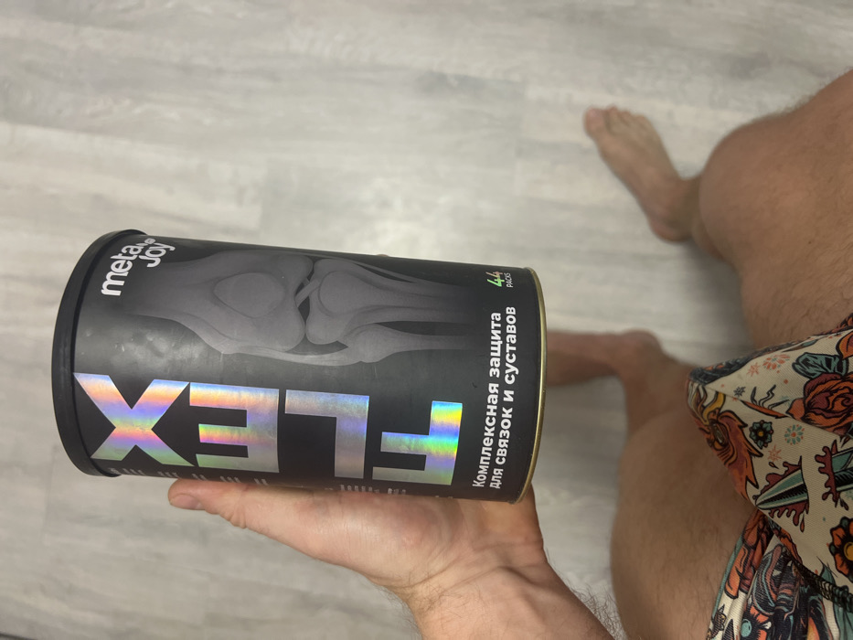 A can of MetaJoy FLEX supplement with a knee joint X-ray graphic is held in a hand. The label features the word "FLEX" in holographic lettering and Russian text describing it as a comprehensive support for ligaments and joints. The can also indicates "44 PACKS". The image includes parts of a person’s leg and feet.