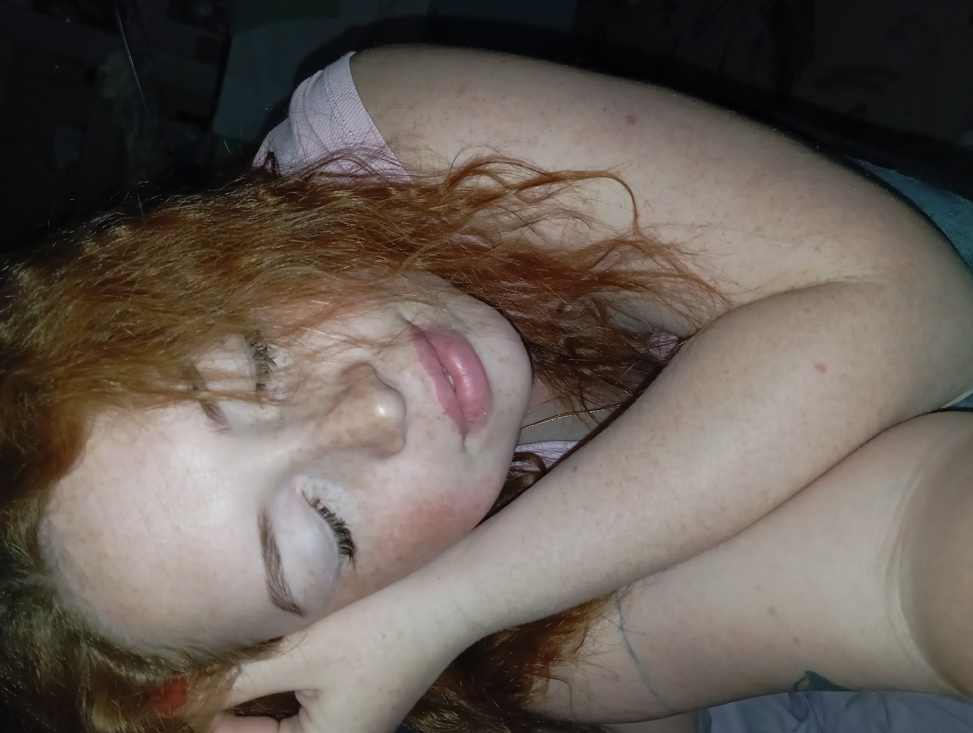 The image shows a person with red hair lying down. Their eyes are closed, and one arm is across their face. The skin is fair with freckles. They are wearing a light pink top. The background is dark and indistinct. There's no indication of the location being Yurgamysh, Russia.