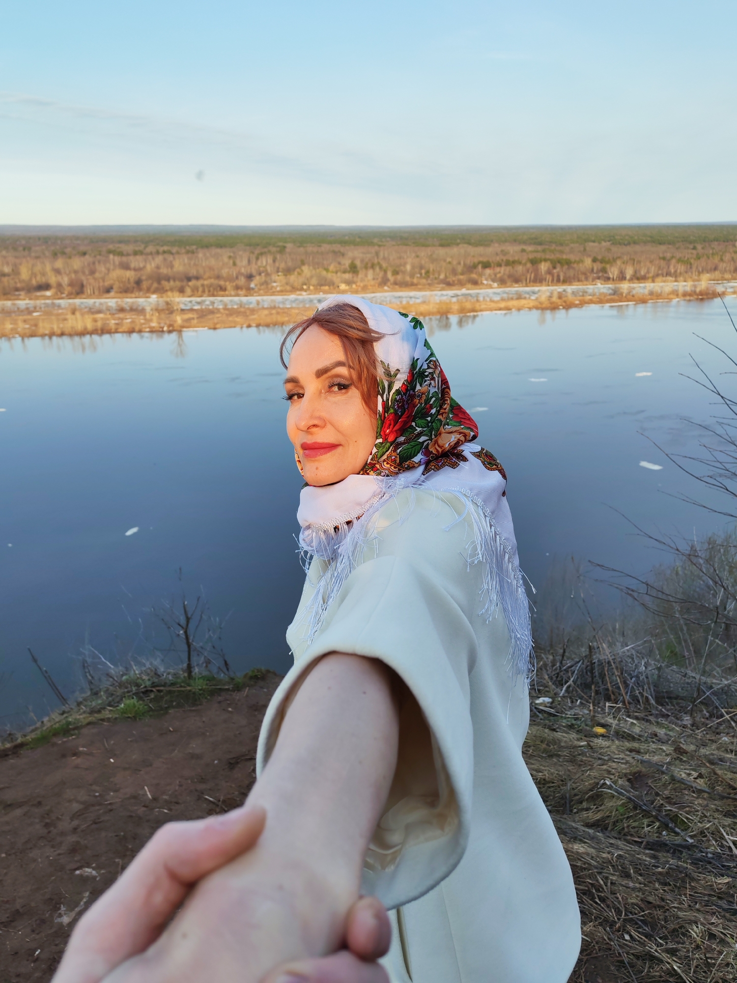 A woman in a white jacket and a colorful headscarf stands on a grassy hill overlooking a river. She is looking at the viewer, offering a hand. The background shows a river, bare trees, and a distant horizon. The scene is possibly located in Sarapul, Russia, based on the style of the headscarf and the landscape.