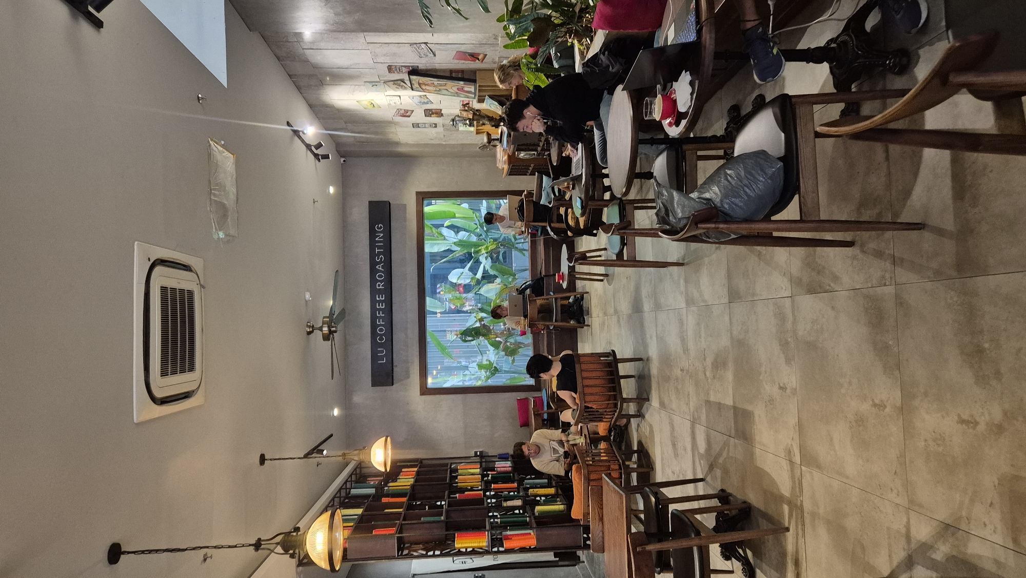 The image depicts the interior of "LU COFFEE ROASTING," a coffee shop located in Đà Nẵng, Vietnam. The environment is that of a contemporary cafe, characterized by a mix of modern and rustic elements.
The space features a large window with lush greenery visible outside, suggesting an urban setting with a touch of nature. Inside, several patrons are seated at various tables, engaged in activities such as working on laptops or conversing.
Objects present include wooden tables and chairs, a prominent bookshelf filled with colorful items, decorative wall art, a ceiling fan, and lighting fixtures. The floor is tiled with large, light-colored square tiles. The overall ambiance suggests a relaxed and studious atmosphere, common in coffee shops that cater to students and professionals.
Specific individuals observed include:
- A person seated at a table on the left, looking down.
- Another person seated at a table further back, facing left.
- Two individuals at a table in the middle ground, one with a laptop.
- A person in the foreground seated at a table, with a laptop and a drink.
- Another person seated to the right of the previous individual, also with a laptop.
- A person in the background on the far right, seated at a table with a laptop.
The lighting appears to be a combination of ambient interior lighting and natural light filtering through the large window. The color palette is muted, with browns, grays, and the vibrant colors from the bookshelf providing accents. The scene captures a typical day in a bustling coffee shop, with people utilizing the space for work, relaxation, and social interaction.