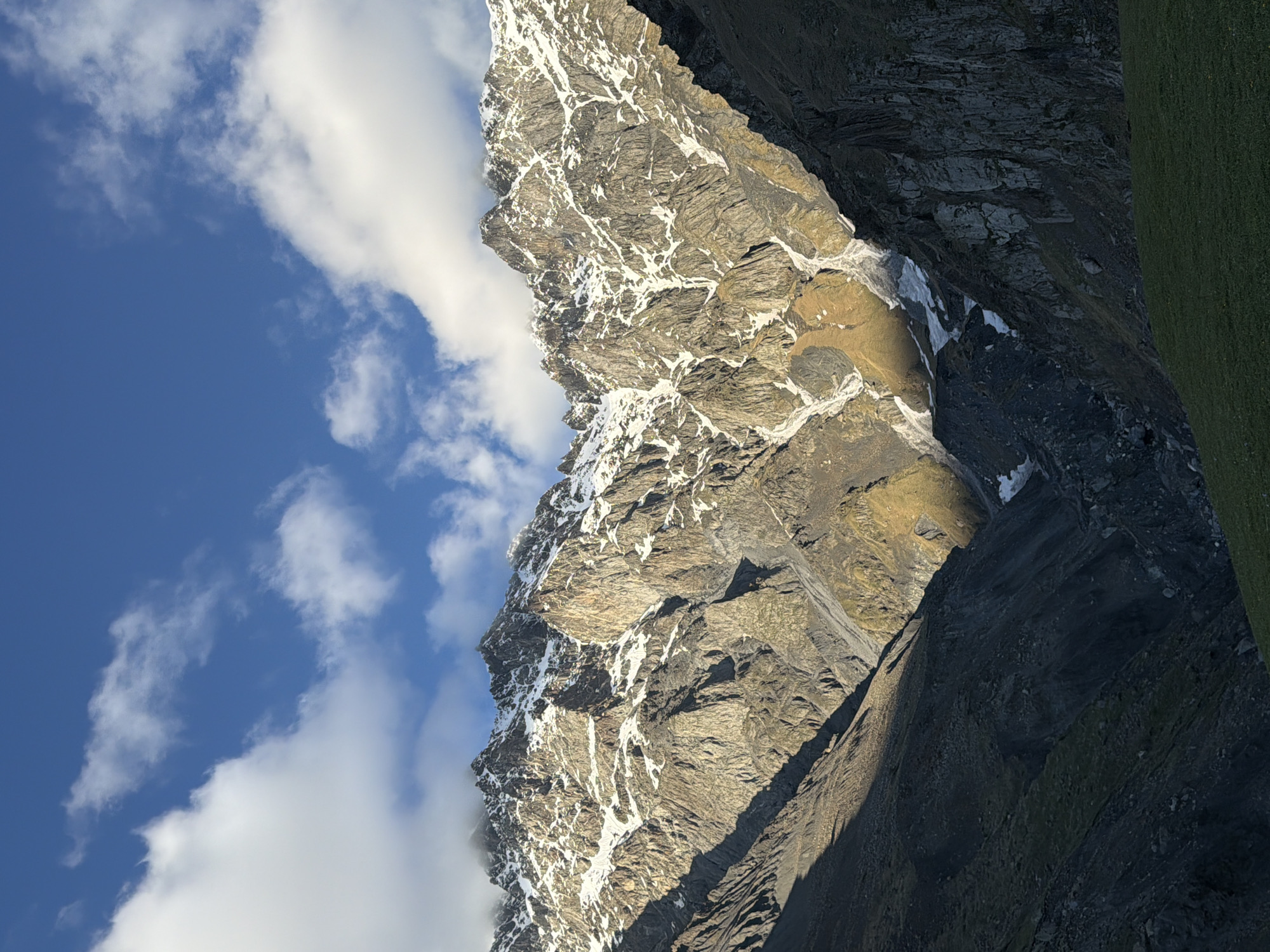 The image shows a steep mountain ridge in Stepantsminda, Georgia. The ridge is composed of gray rock with patches of snow clinging to the crevices and higher elevations. Blue sky with white clouds is visible behind the ridge. Adjacent to the snow-covered ridge is a darker, rocky slope. A patch of green grass can be seen on the right side of the image.
